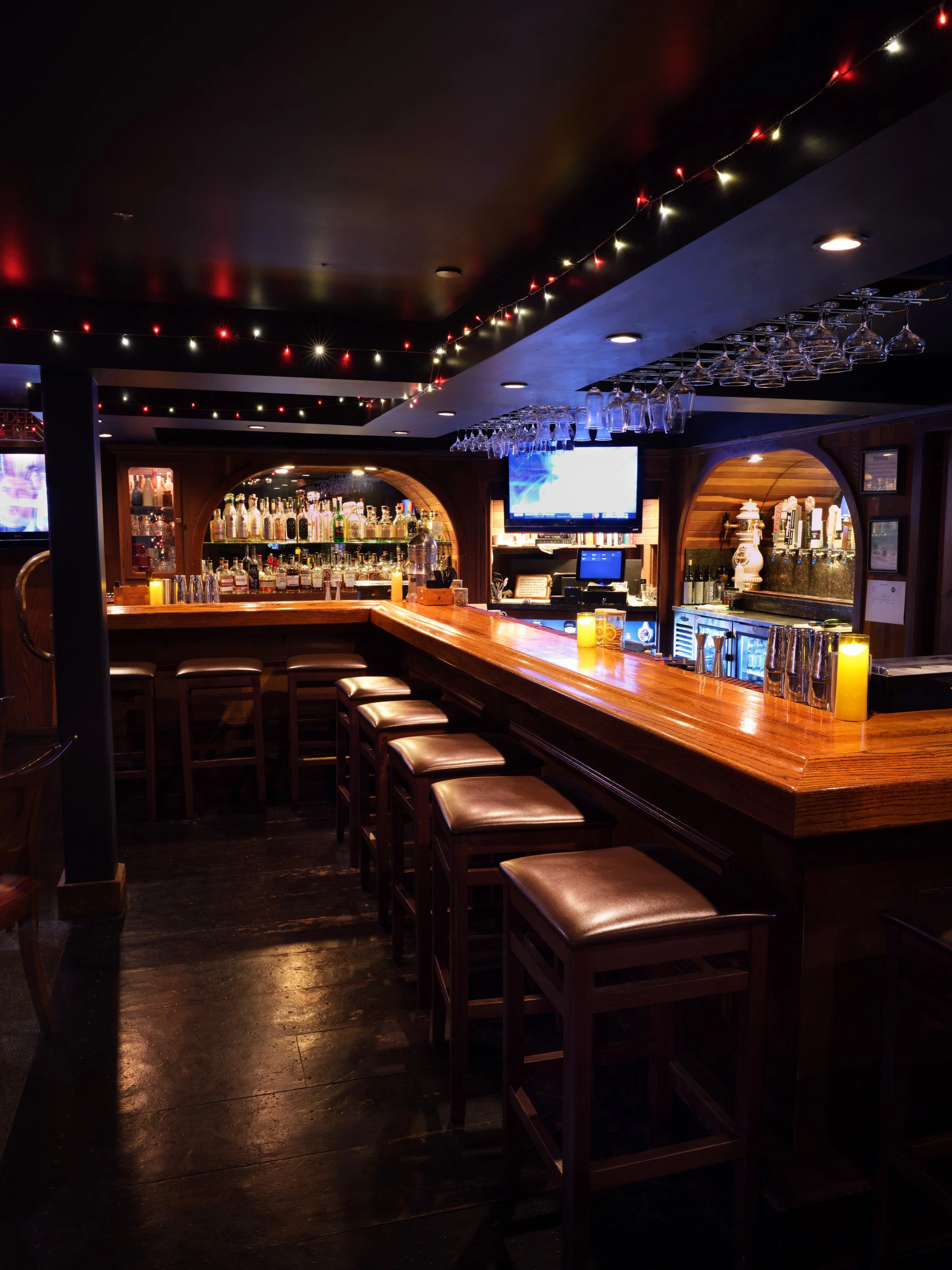A dimly lit bar with a wooden counter, bar stools, shelves of liquor bottles, hanging wine glasses, and a mounted TV screen.