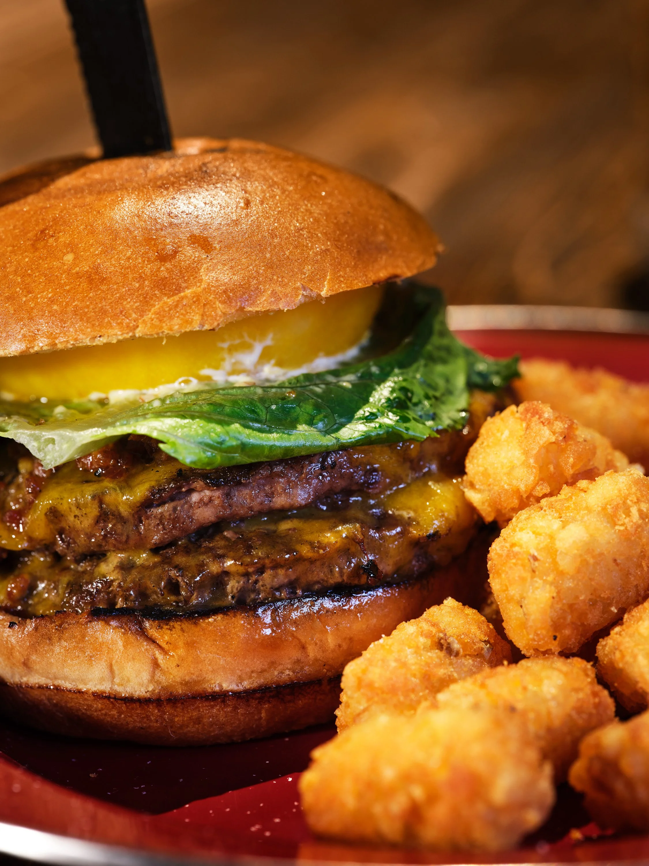 Close-up of a cheeseburger with lettuce, pickles, and melted cheese, alongside crispy tater tots on a plate.