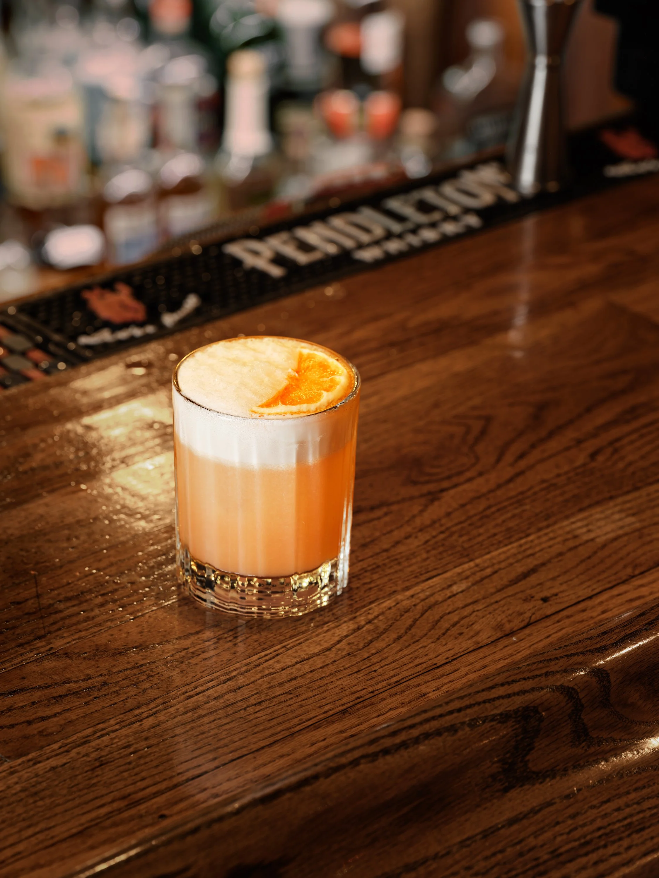 A tall glass of a layered orange and white cocktail garnished with an orange slice on a wooden bar counter.
