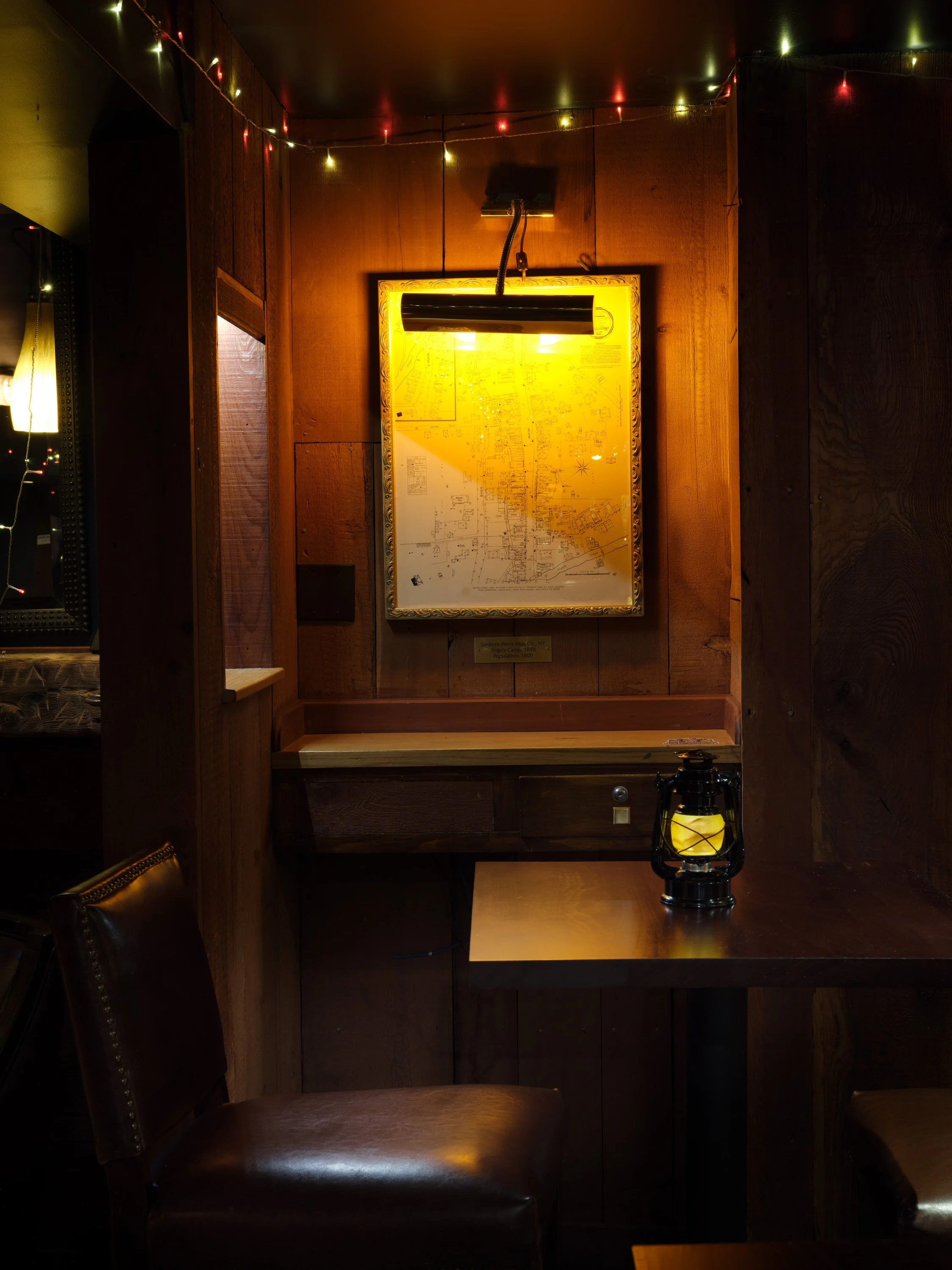 A wall with a framed map or diagram illuminated by a yellow lamp, surrounded by dark wood paneling, string lights, and a lantern on a table below.