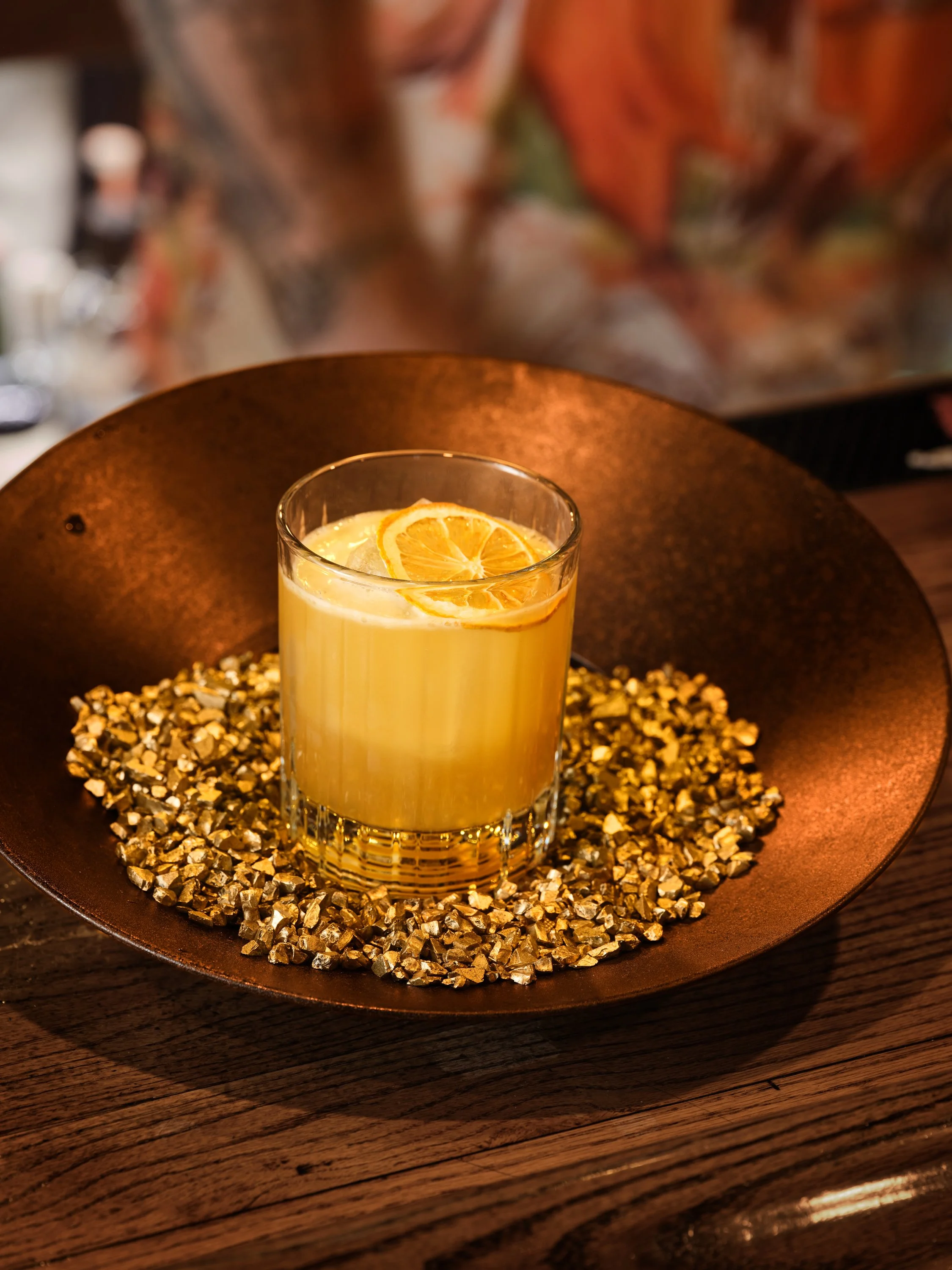 A cocktail with a lemon peel garnishing it, placed on a gold pan with gold-colored stones, on a wooden table.