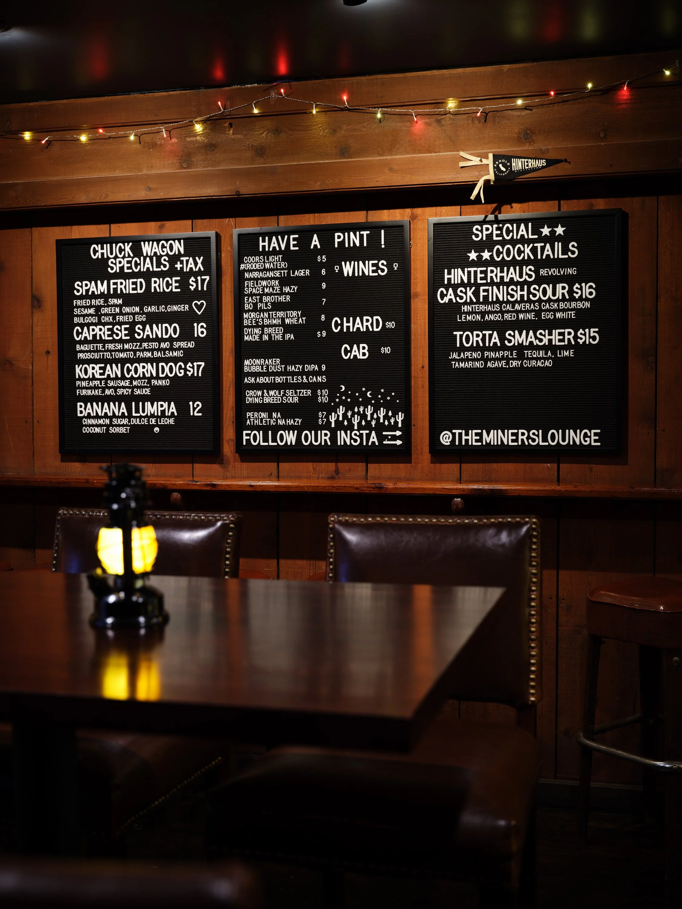 Wood-paneled wall with string lights, three black menu boards displaying food and drink options, and a small decorative pennant with 'Hinterhaus' on a shelf. In the foreground, a dark wooden table with a small lantern, empty chairs around, and a dark