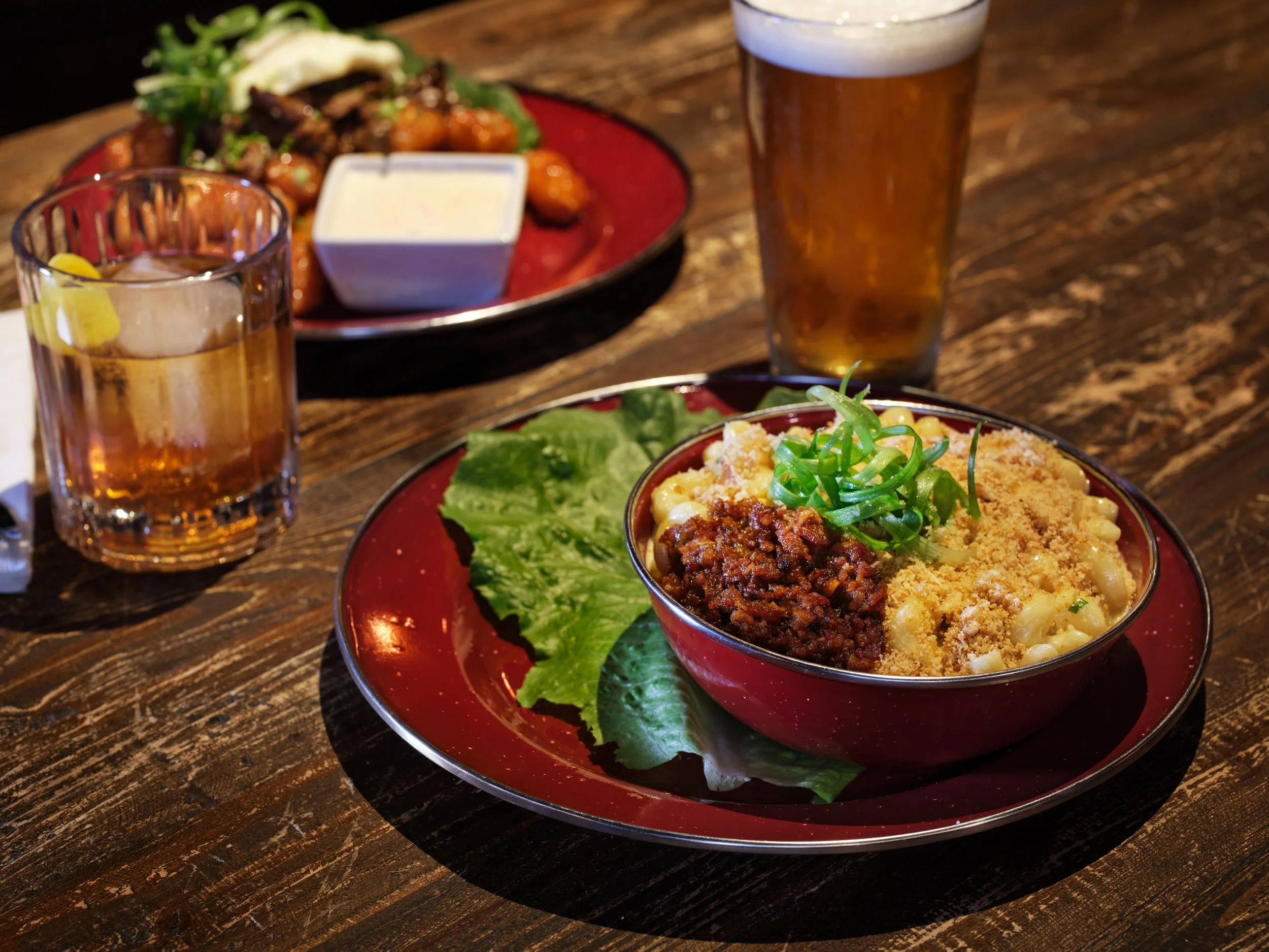 A bowl of macaroni and cheese topped with chopped green onions, on a rustic wooden table. In the background, there is a sticky tots with green onion garnish and a small container of dipping sauce, and two glasses of beer.