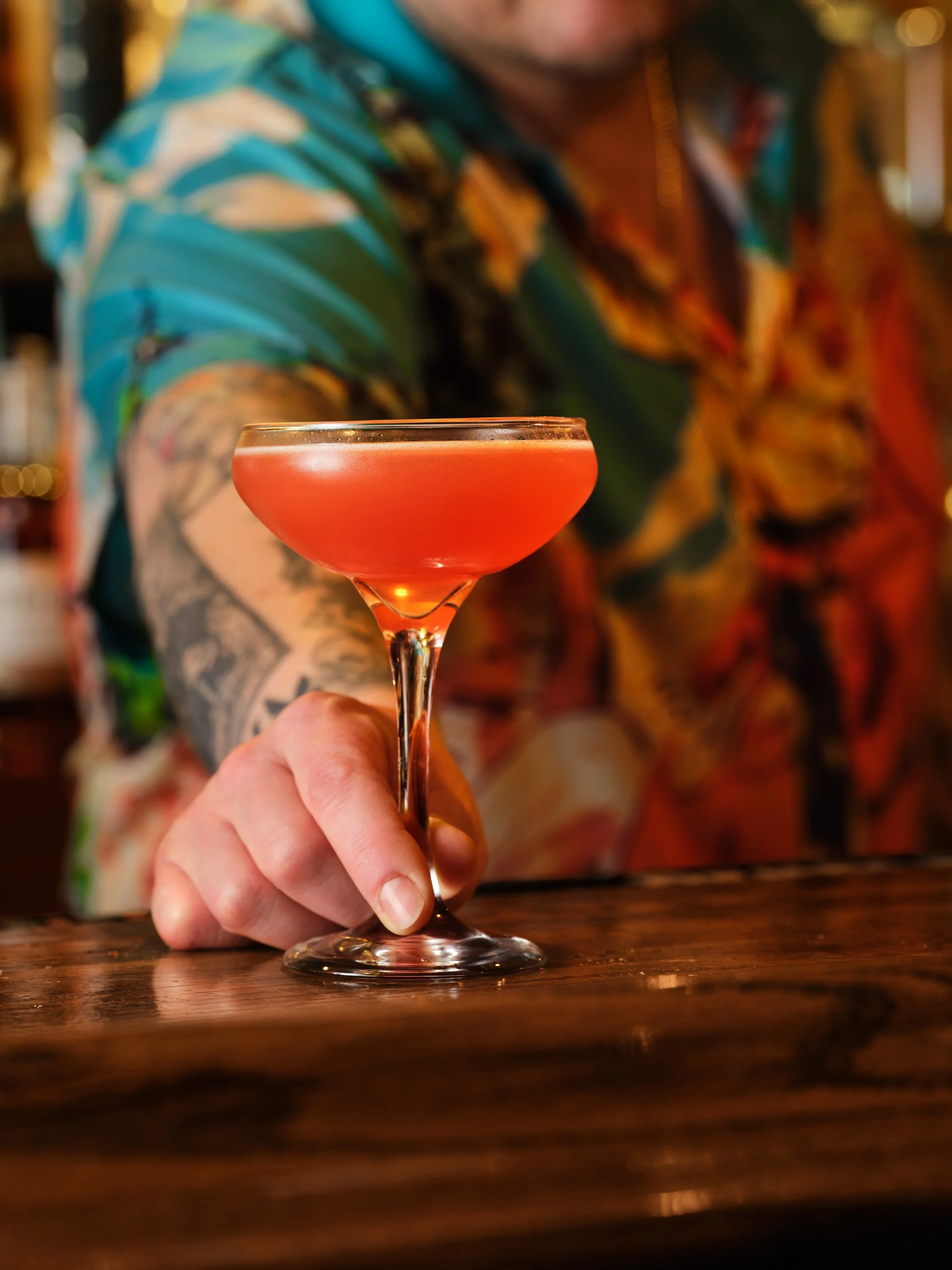 A person holding a pink cocktail in a coupe glass, with a blurred background, wearing a colorful, floral shirt.