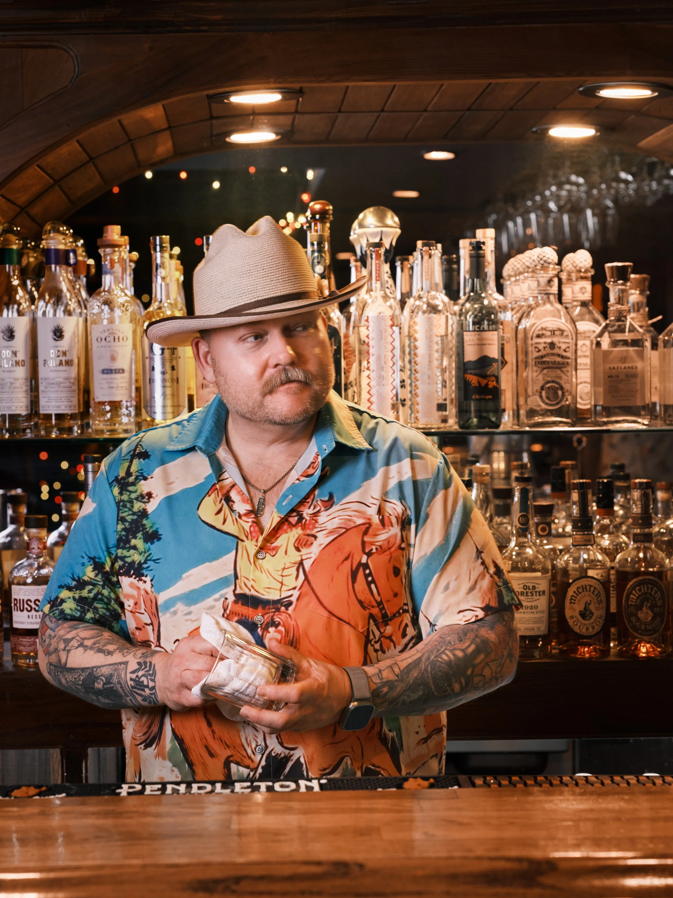 A man with tattoos on his arms wearing a beige fedora and a colorful Hawaiian shirt behind a bar, holding a cocktail glass, with liquor bottles on shelves in the background.