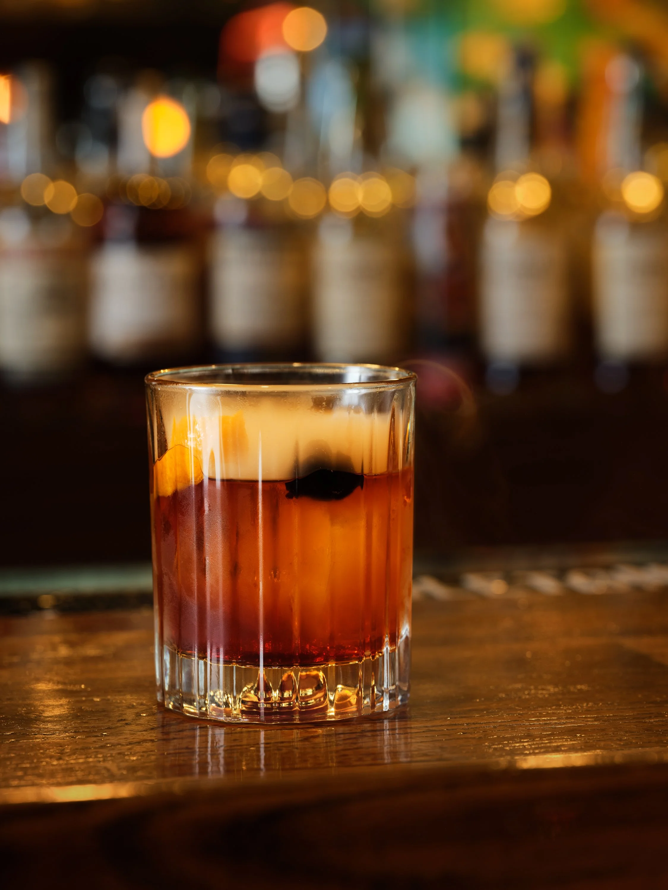 Close-up of a glass of amber-colored liquor on a wooden bar counter with blurred bokeh lights in the background.