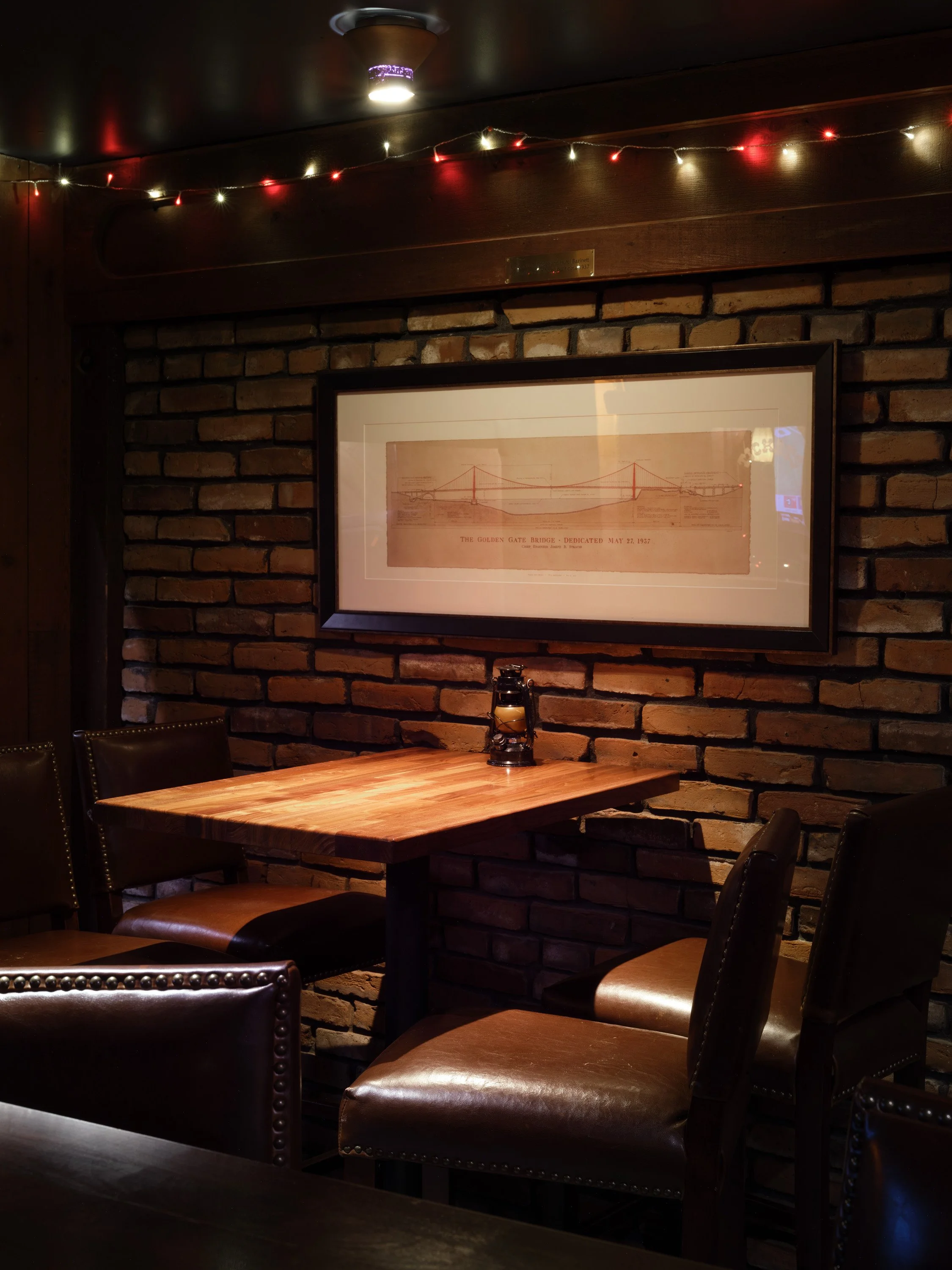 A cozy indoor setting with a brick wall, wooden table, leather chairs, framed artwork, and string lights overhead.