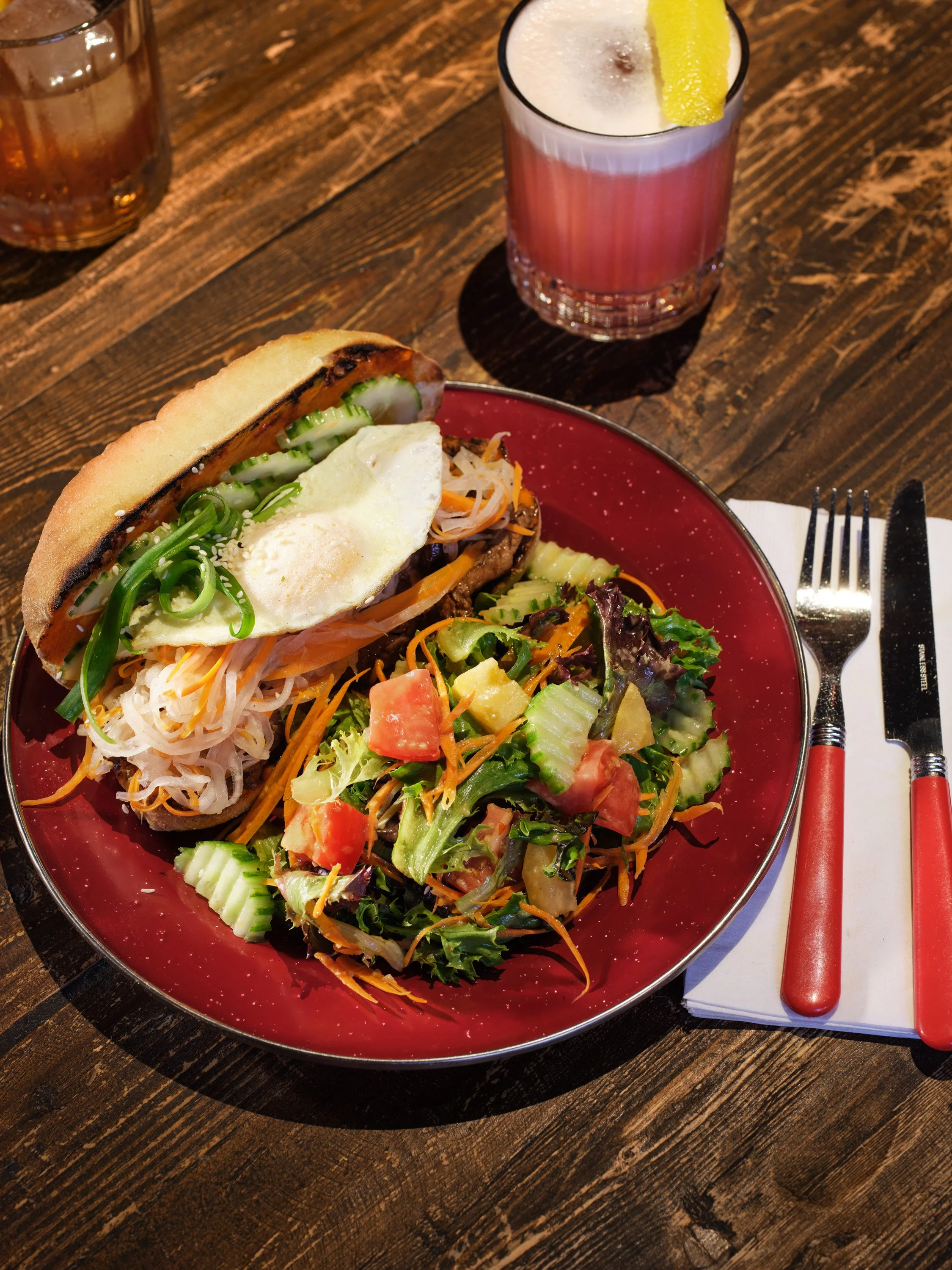 A plate with a bulgogi banh mi sandwich topped with a fried egg, served with a salad featuring mixed greens, cucumber, tomato, shredded carrots and croutons.