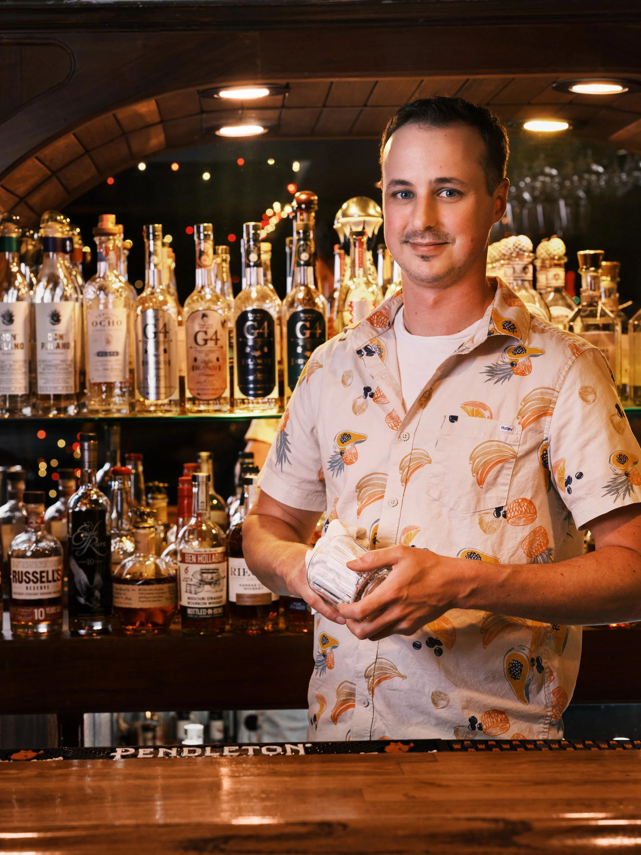 A man with short dark hair wearing a cream-colored shirt with a tropical fruit pattern, stands behind a bar with bottles of liquor in the background. He is smiling slightly and holding a cocktail glass.