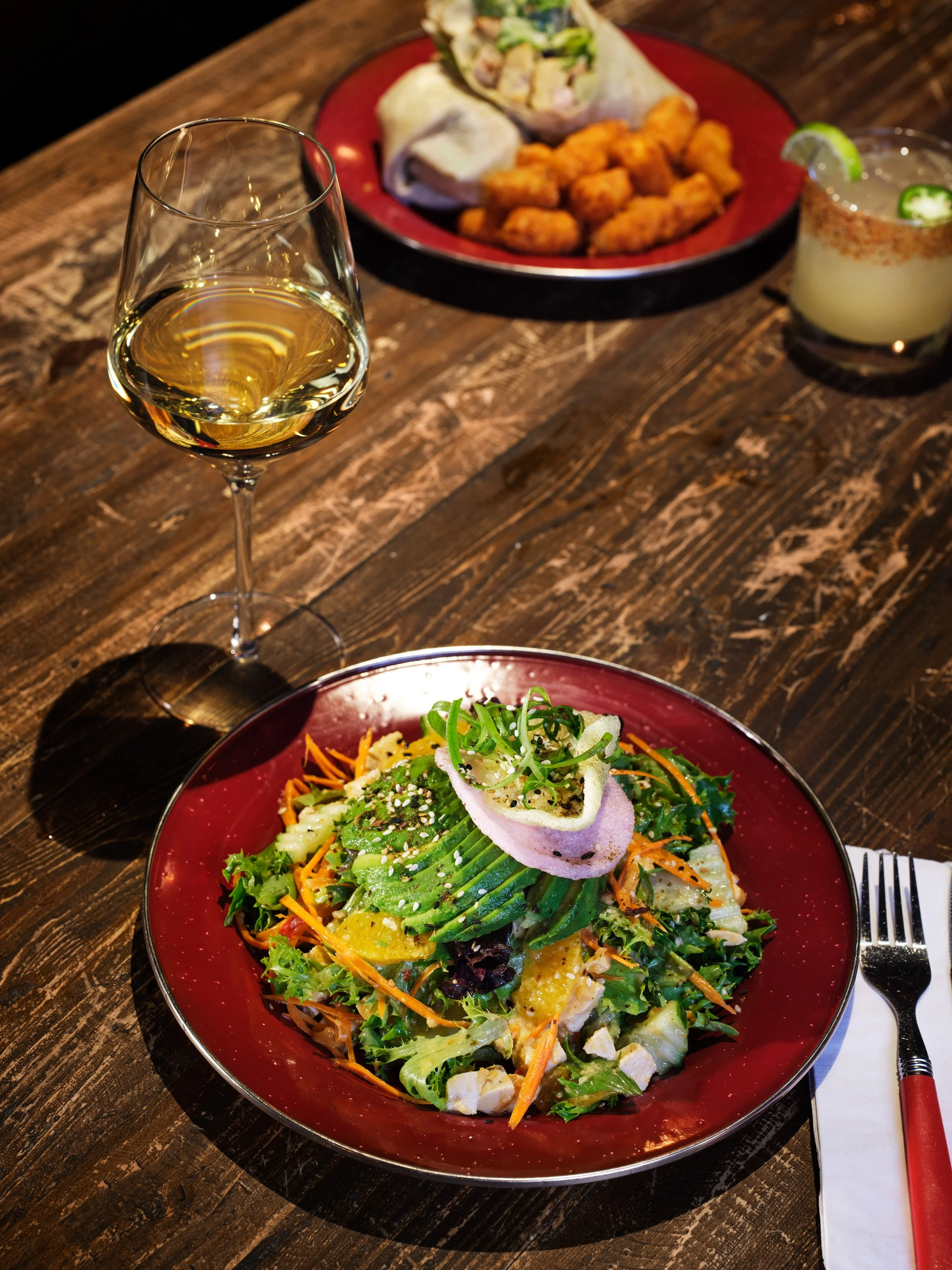 A colorful salad with avocado, carrots, and greens in a red bowl, with a glass of white wine and a plate with spring rolls, fried chicken bites, and a cocktail with lime and jalapeño slices on a wooden table.