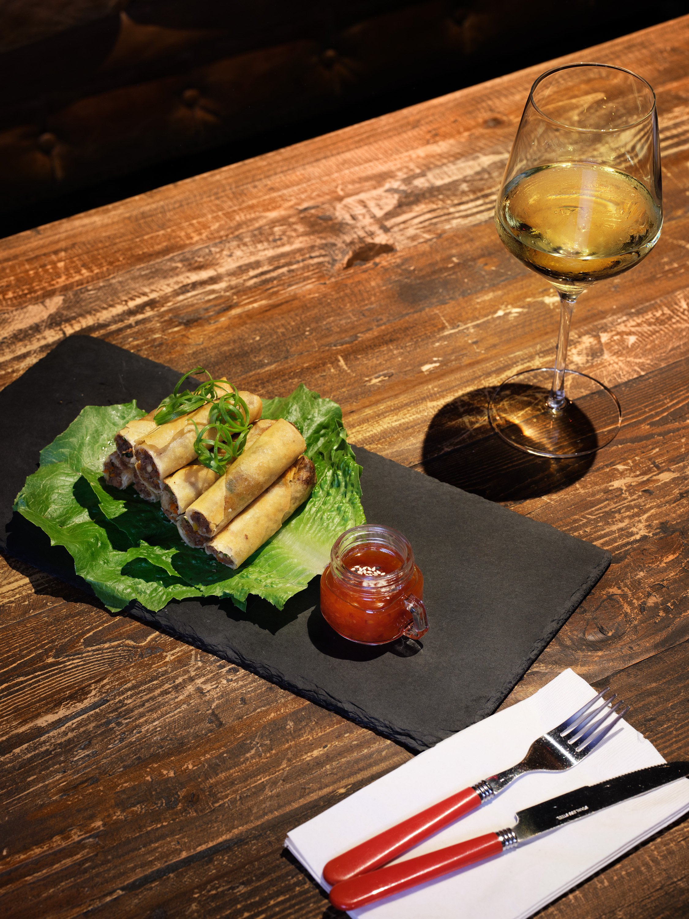 A wooden table with a black slate serving platter holding lumpia garnished with green herbs on lettuce leaves, a small jar of dipping sauce, a wine glass of white wine, and a set of red-handled fork and knife on a white napkin.