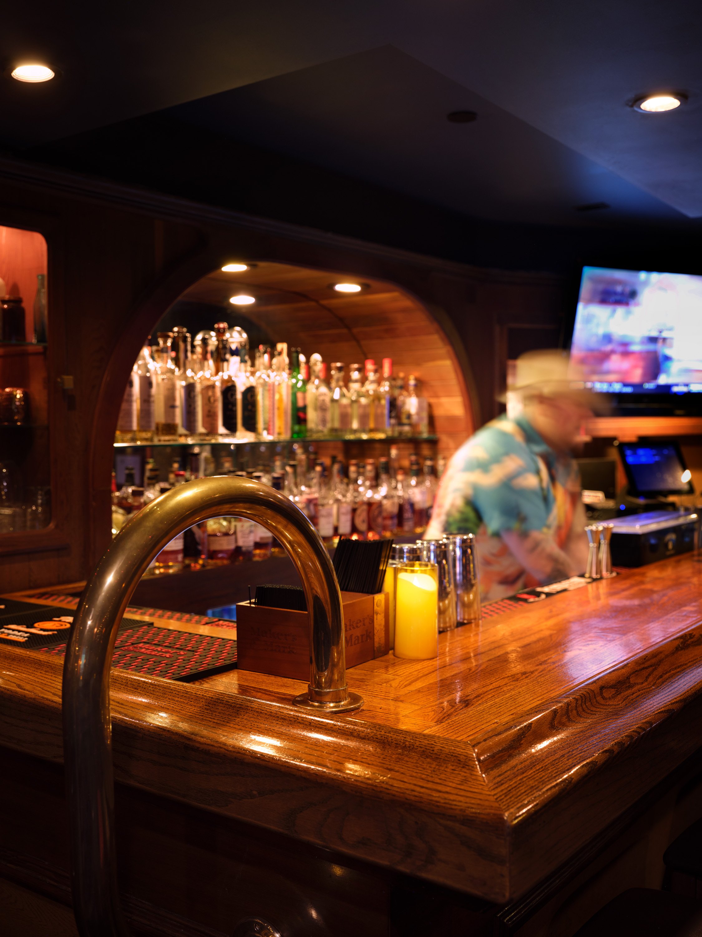 A wooden bar counter in a dimly lit bar with bottles of alcohol in the background, a bartender wearing a colorful shirt, and a television screen on the wall.