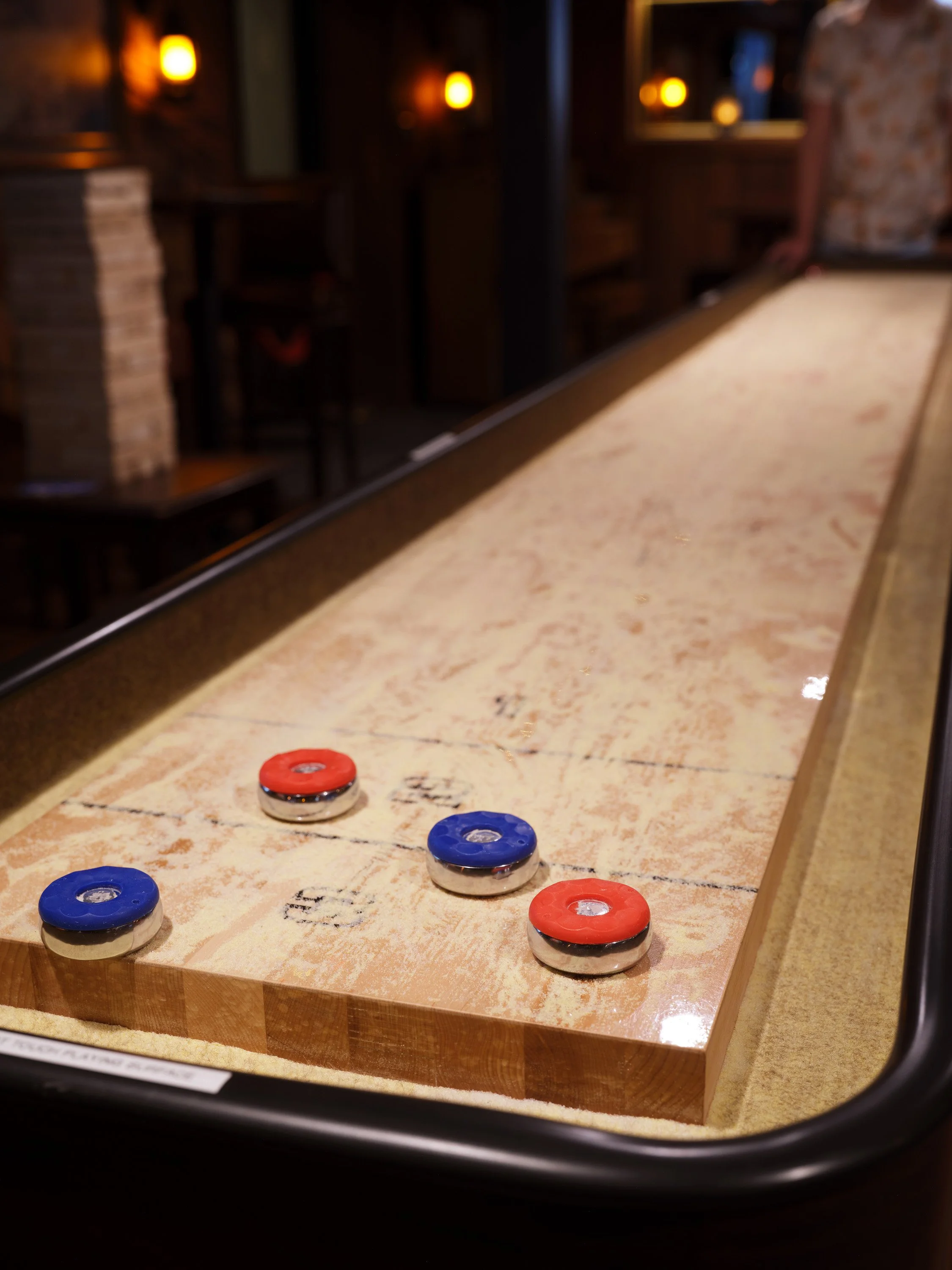 A shuffleboard game with red and blue pucks on a wooden surface in a dimly lit indoor setting, possibly a bar or game room.