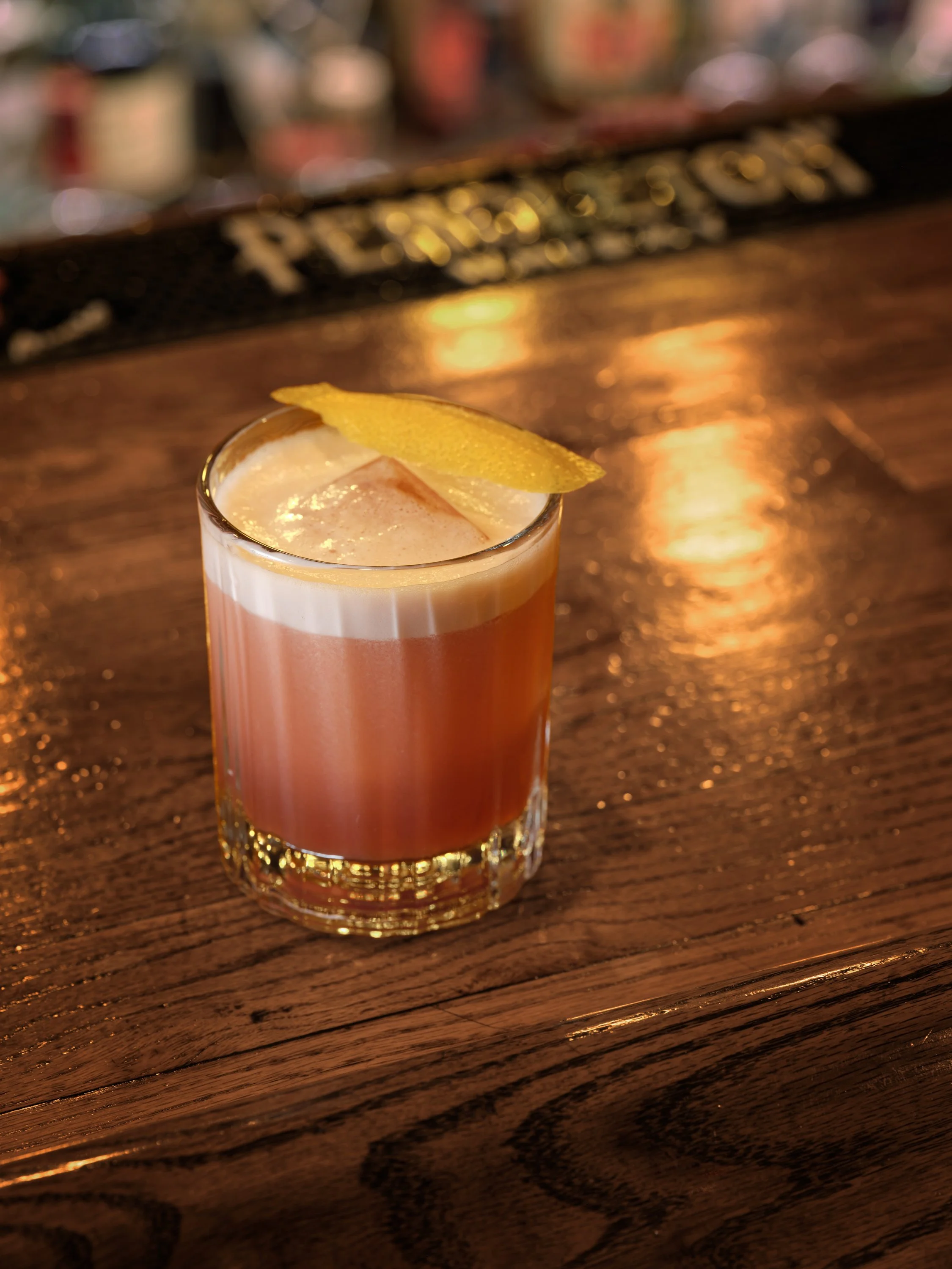 A cocktail with a lemon twist garnish on a wooden bar counter.