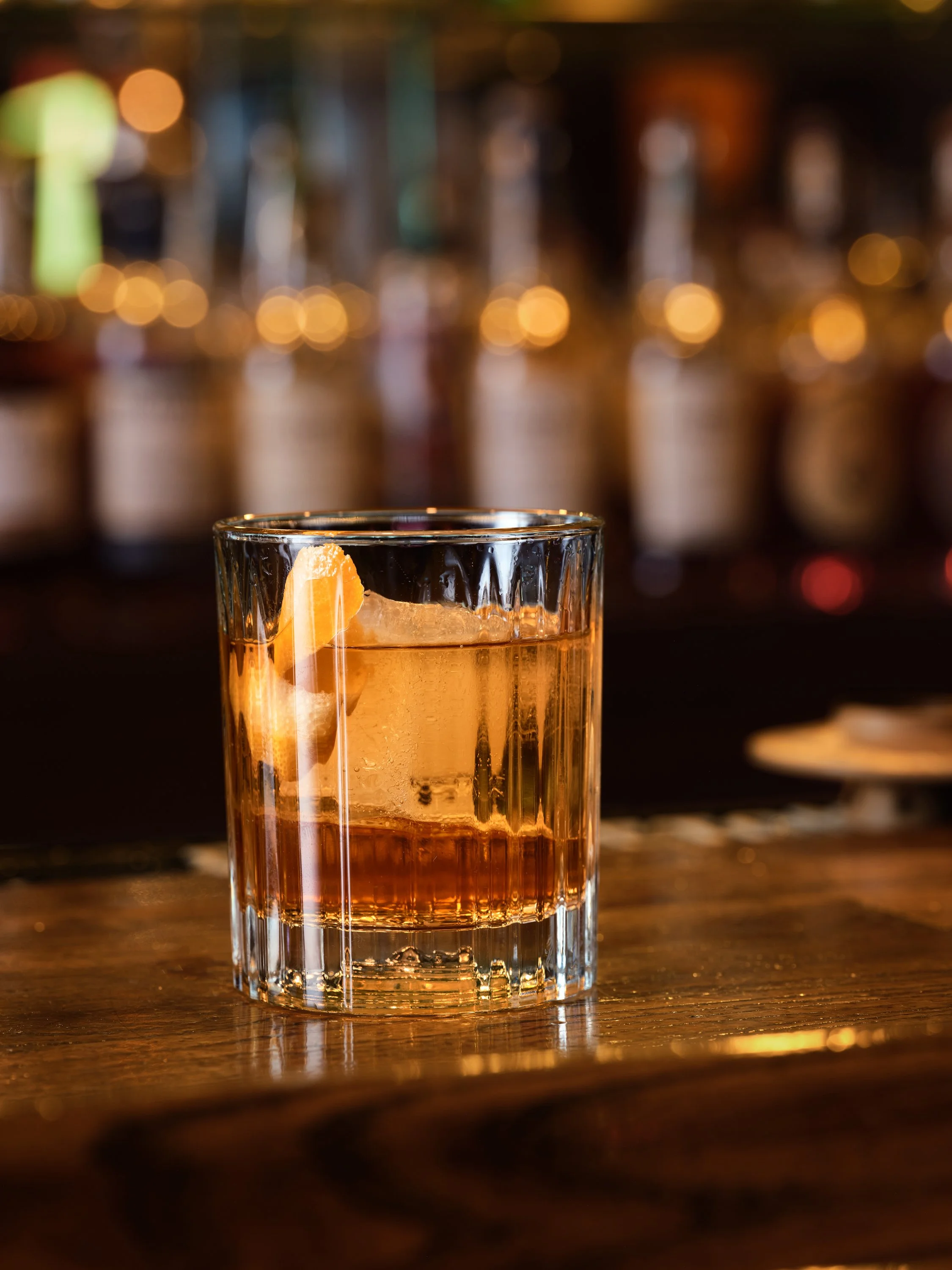 An old fashioned on a large  ice cube on a wooden bar counter, blurred background of bottles and warm lighting.