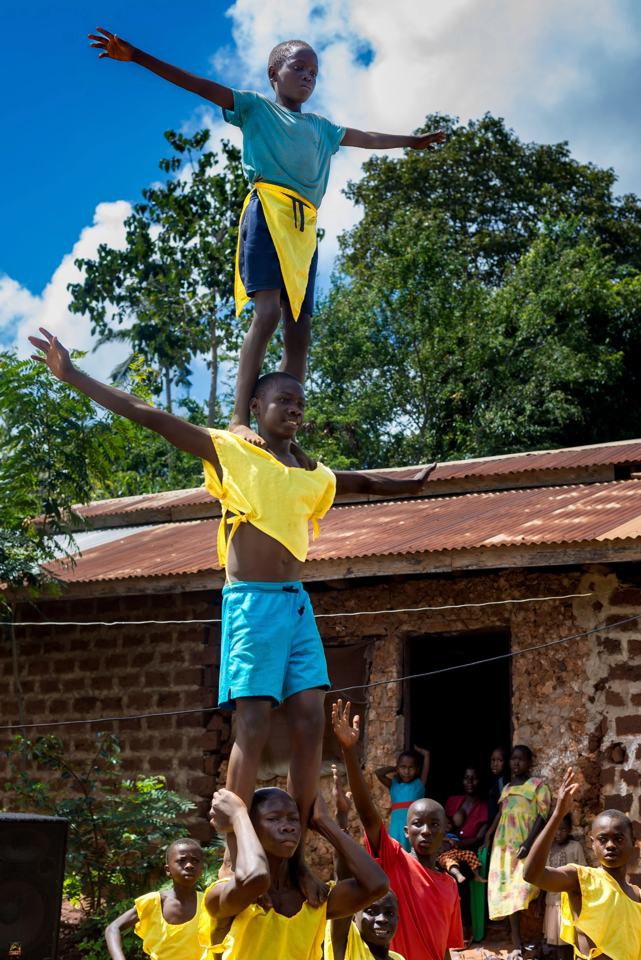 Children performing a human pyramid outdoors, with a brick building and trees in the background.