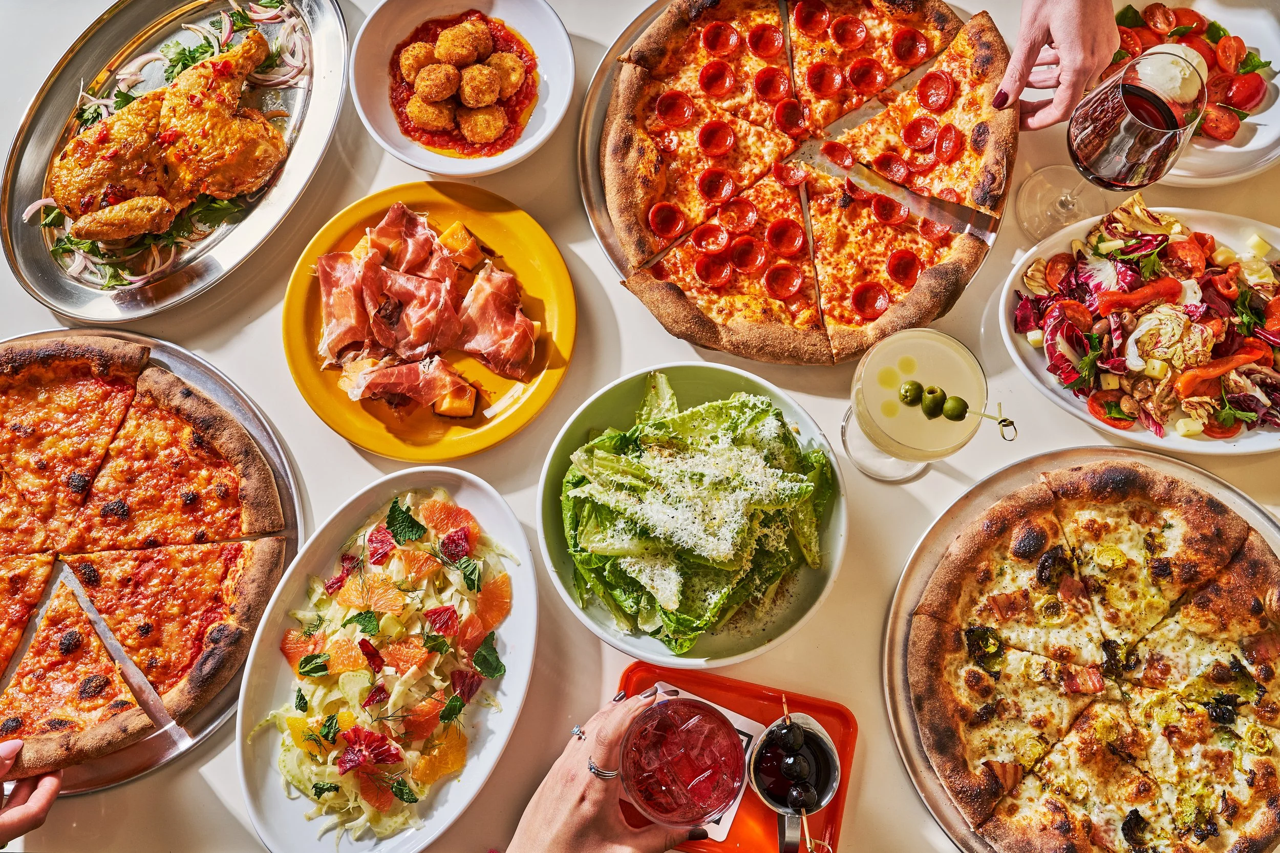 An overhead view of a table filled with various dishes including pepperoni pizza, a white sauce pizza, chicken, prosciutto with melon, mixed salads, arancini, and drinks including cocktails and red wine.