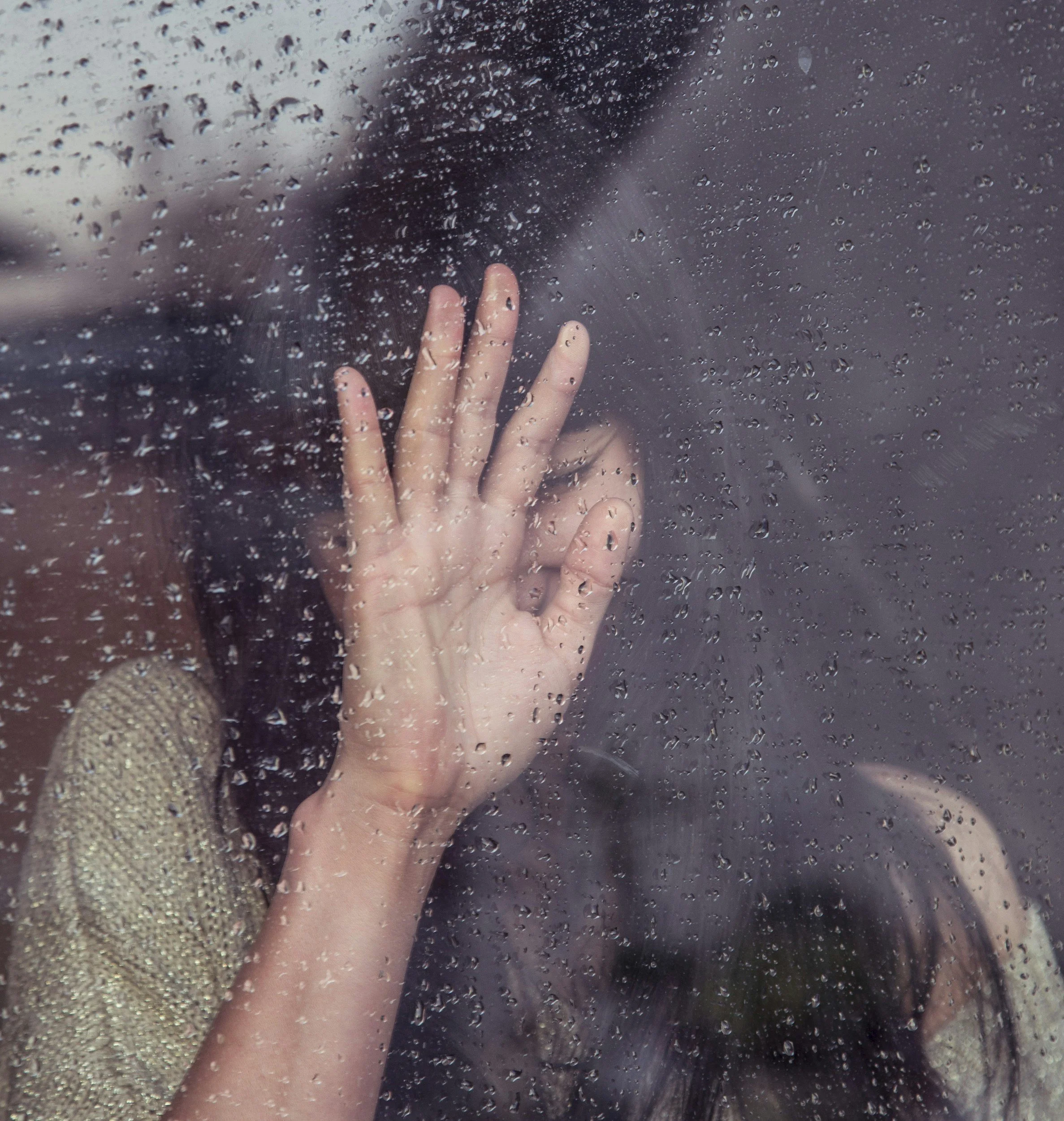 A woman with dark hair and a beige, textured top presses her hand against a rain-covered glass window, obscuring her face.