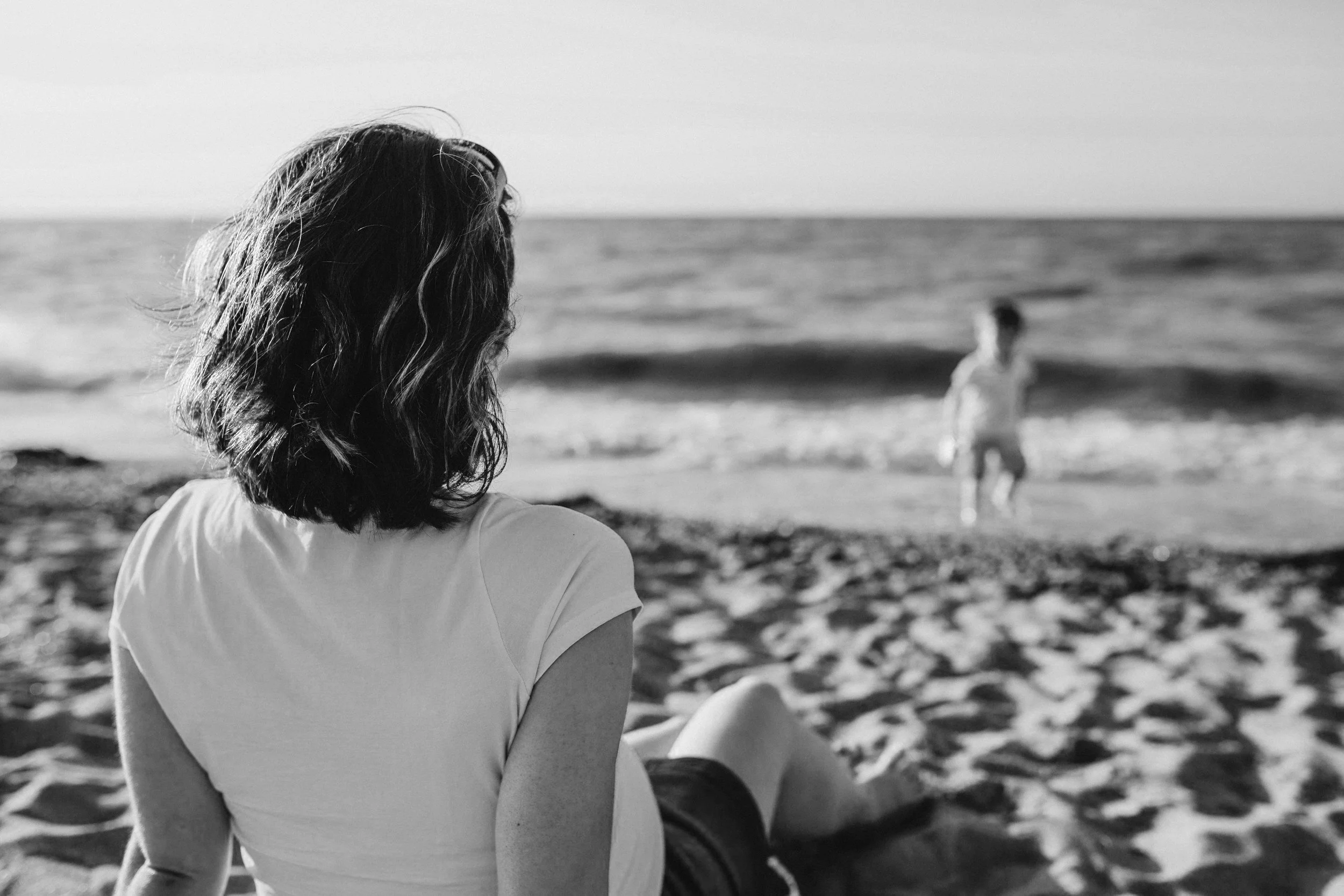 A woman with shoulder-length wavy hair sitting on a sandy beach, watching a child near the ocean.