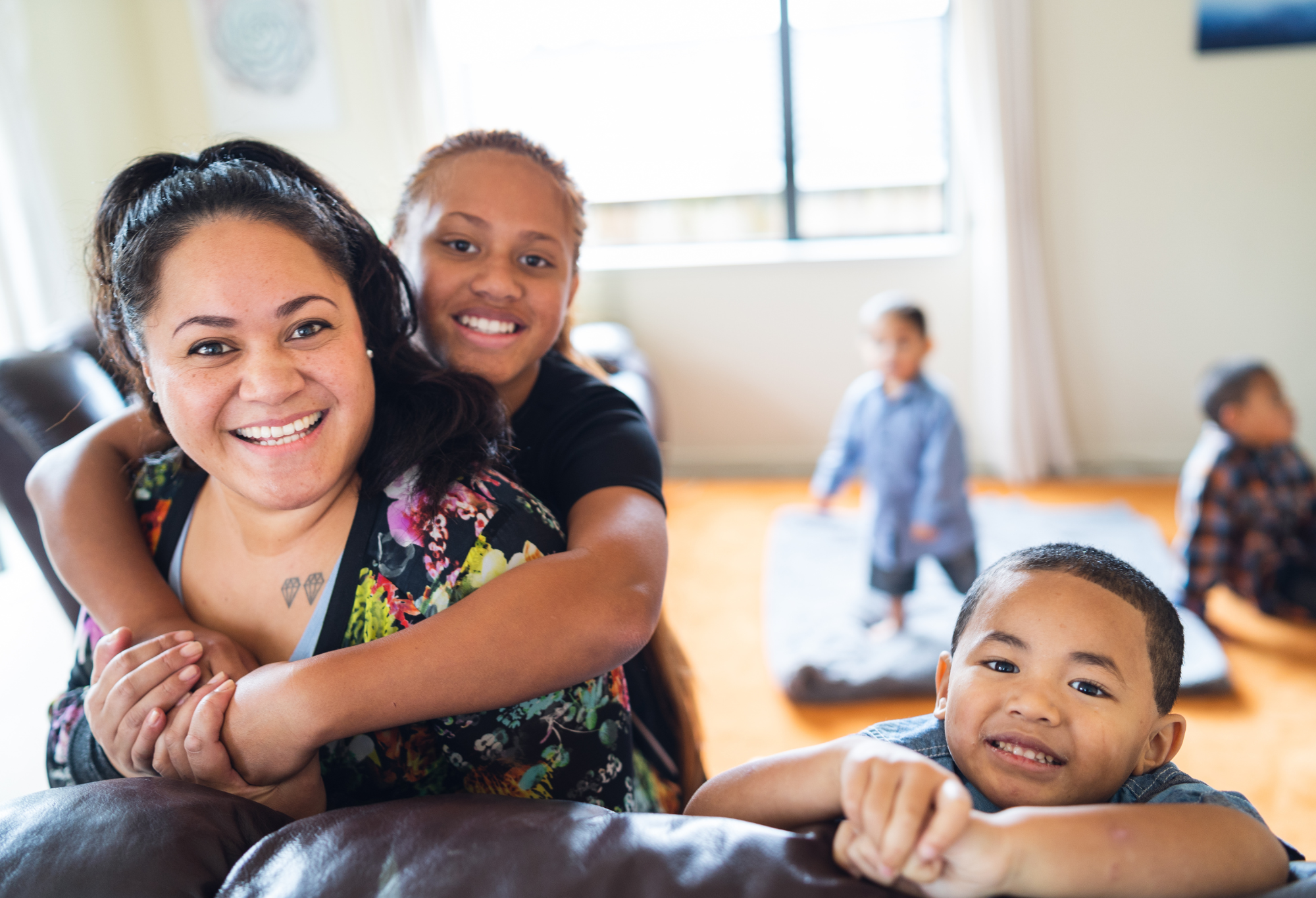 A woman and a girl smiling, with the girl hugging the woman from behind, as three children play in the background on a wooden floor in a bright room.