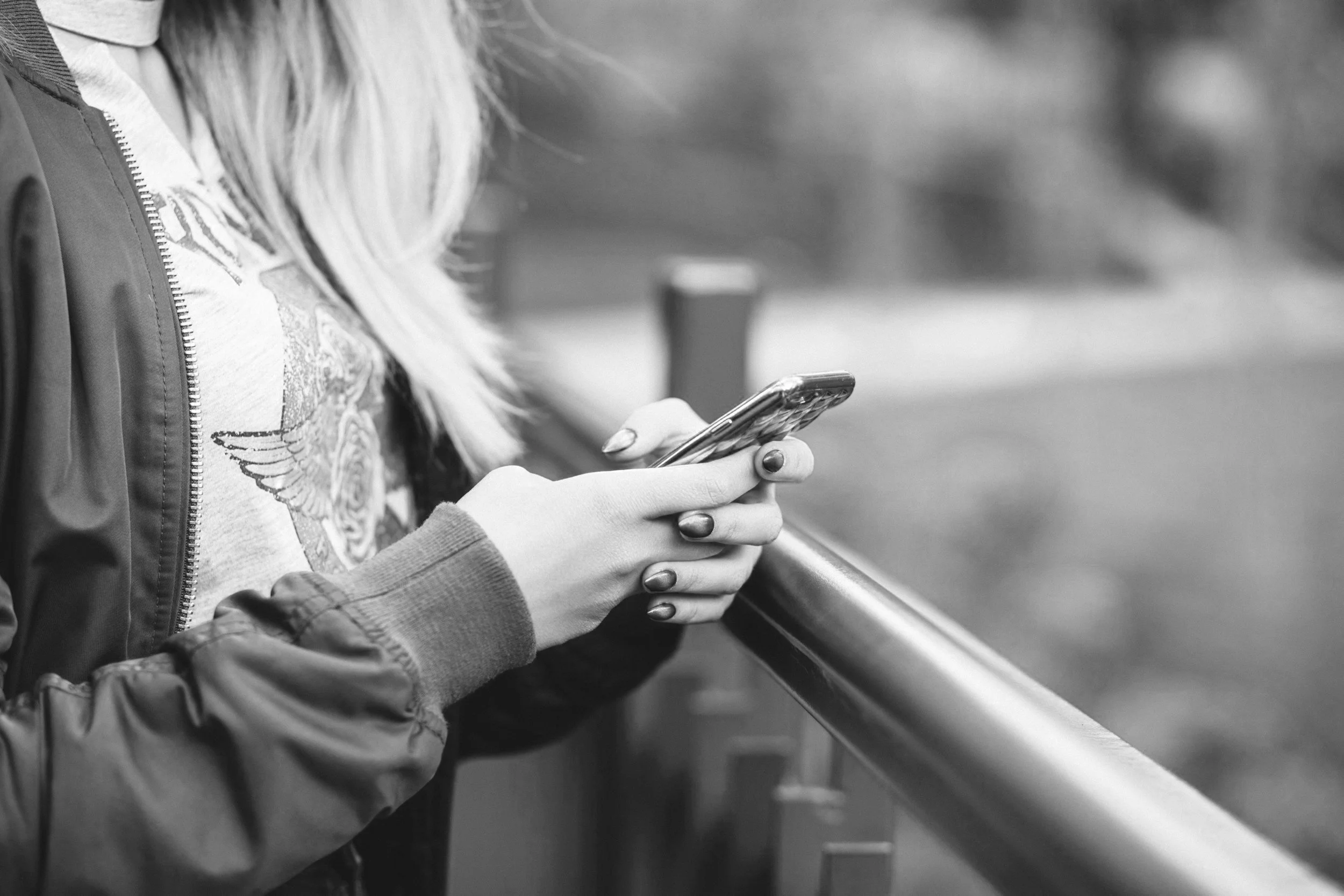 A person with long light-colored hair, wearing a jacket and a graphic t-shirt, is holding and looking at a smartphone while standing outdoors near a metal railing.
