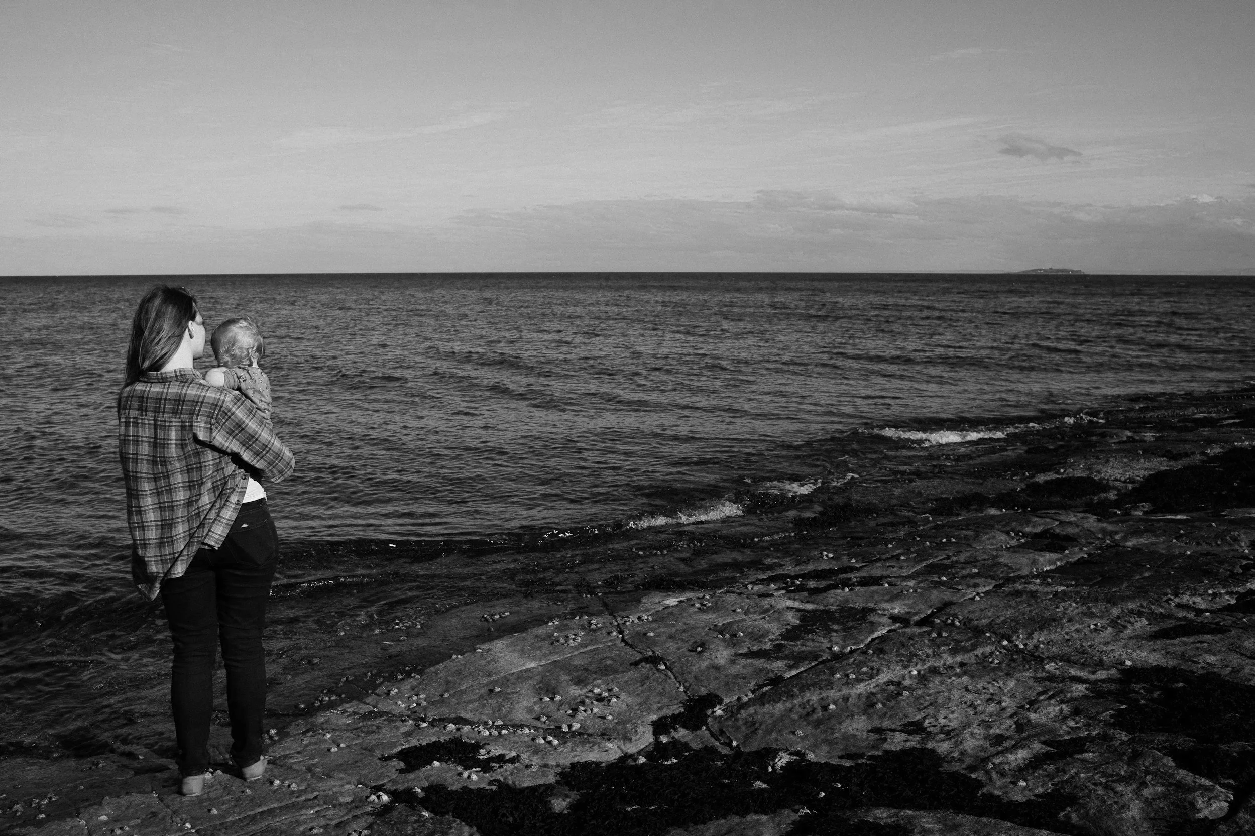 Woman in plaid shirt holding a baby by the rocky shoreline, looking out at the ocean during a cloudy day.