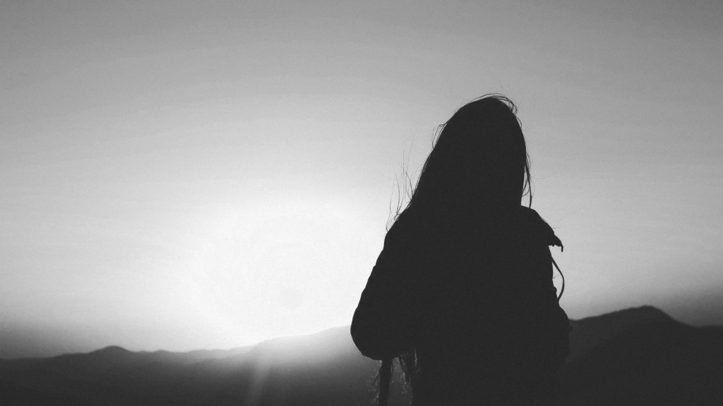 Silhouette of a woman with long hair standing outdoors during sunset or sunrise, with a mountainous landscape in the background.