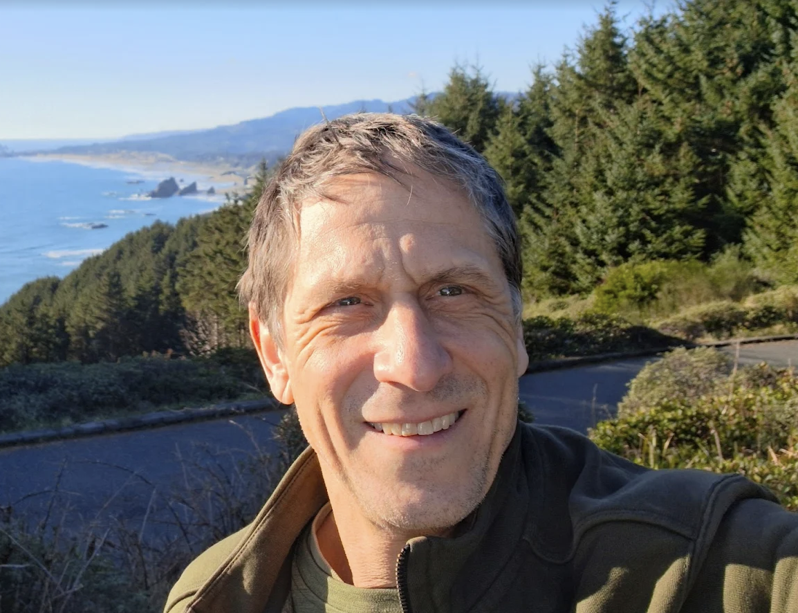 Smiling man taking a selfie outdoors with a scenic coastal view in the background, including trees, water, and hills.