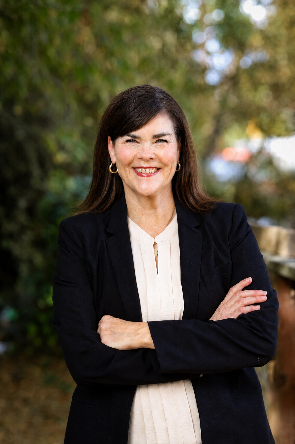 A woman with dark brown hair, wearing a black blazer over a white blouse, smiling with arms crossed outdoors with blurred trees in the background.