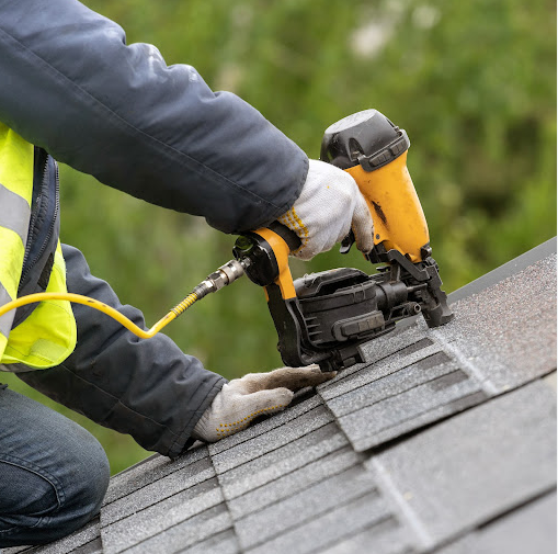 Person using a nail gun to attach shingles to a roof with a safety vest and gloves, outdoors.