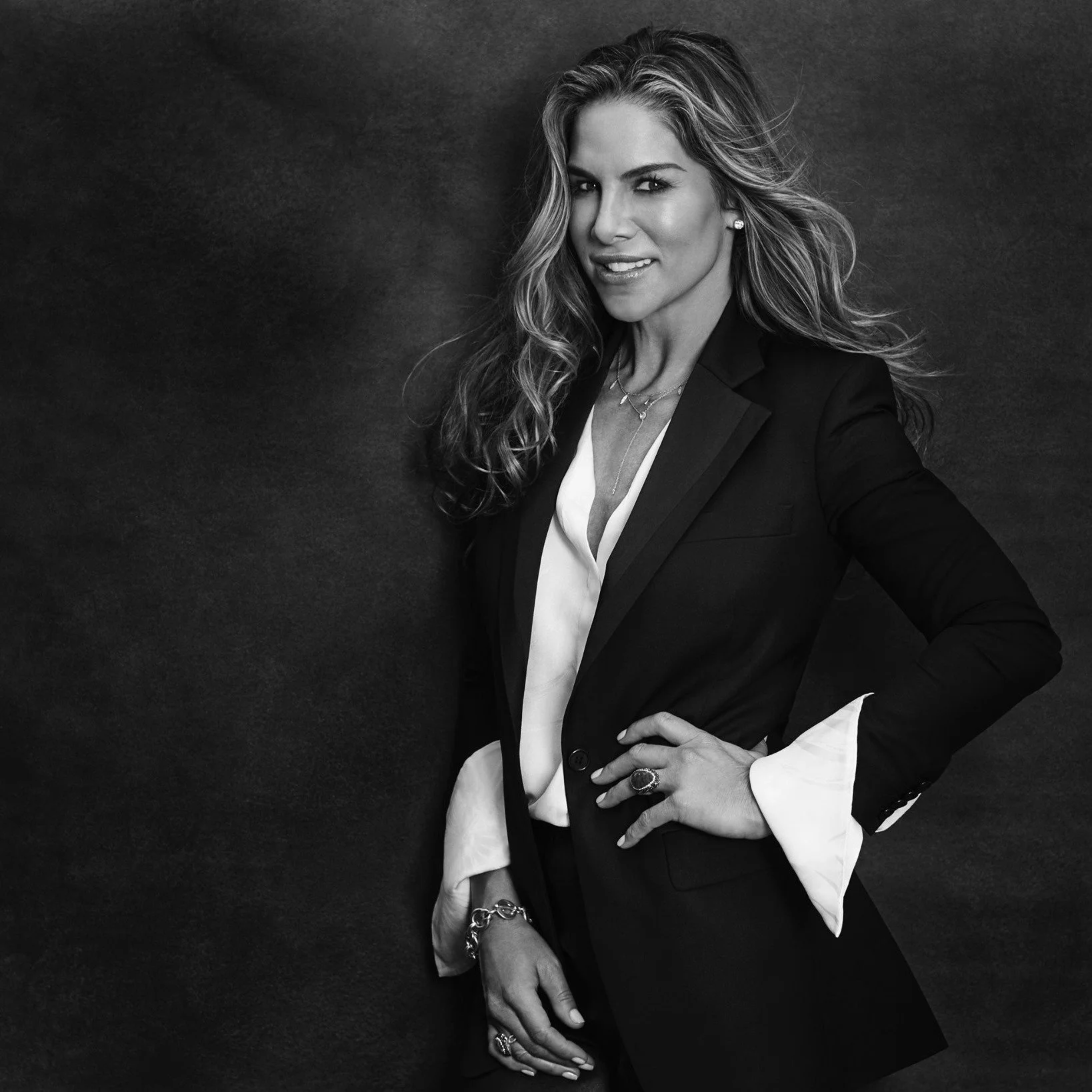 Black and white portrait of a woman with wavy hair, wearing a blazer and jewelry, standing against a dark background, looking at the camera.