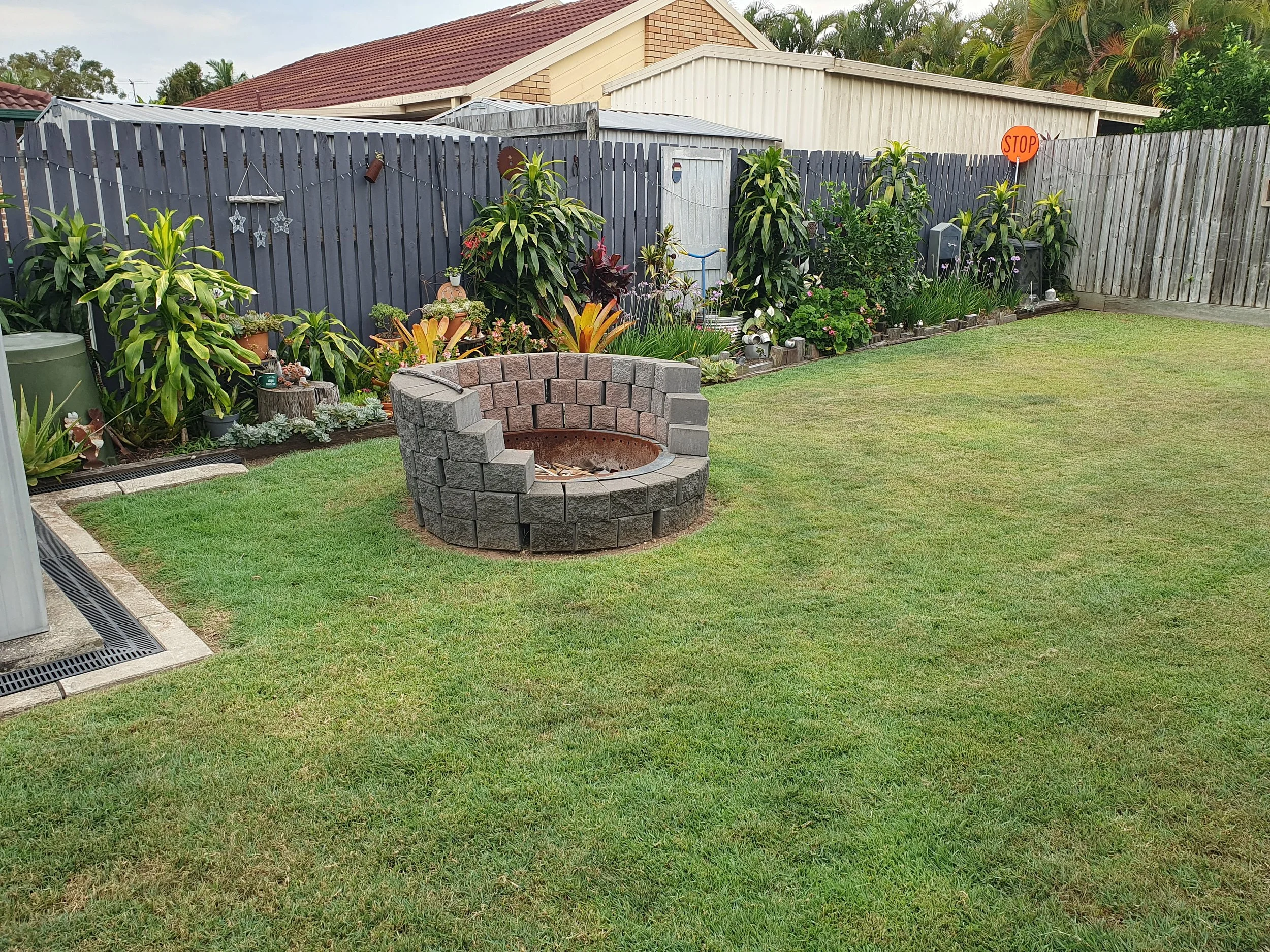 Backyard with fire pit, grass, and garden along a wooden fence.