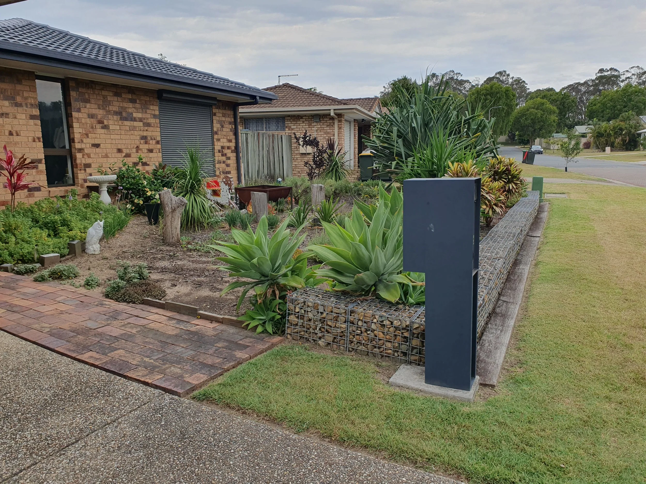 Front yard with brick pathway, garden bed, and window of a brick house; large plants and greenery visible.