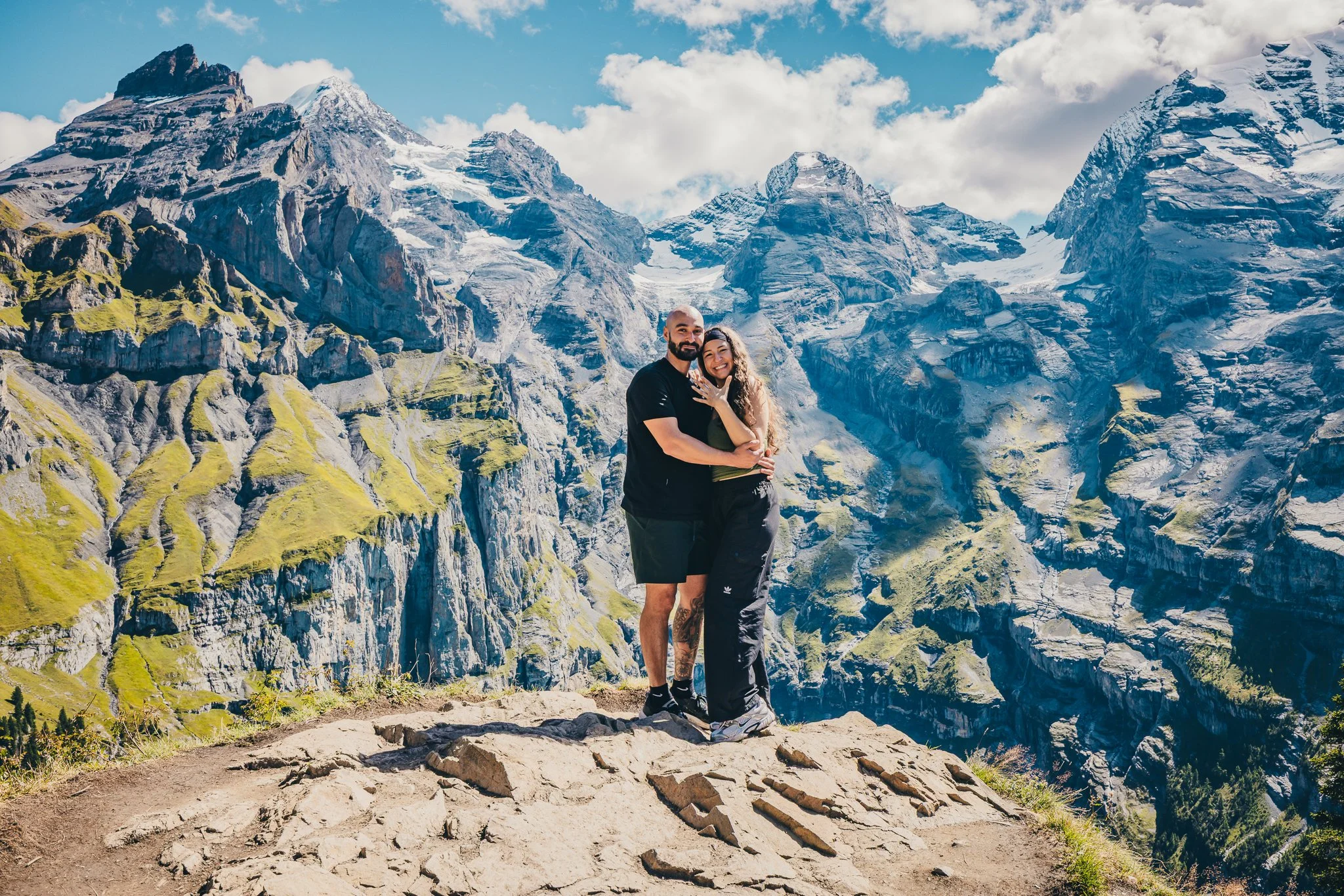 A couple hugging on a rocky ledge with a mountainous landscape featuring snow-capped peaks and green slopes in the background.