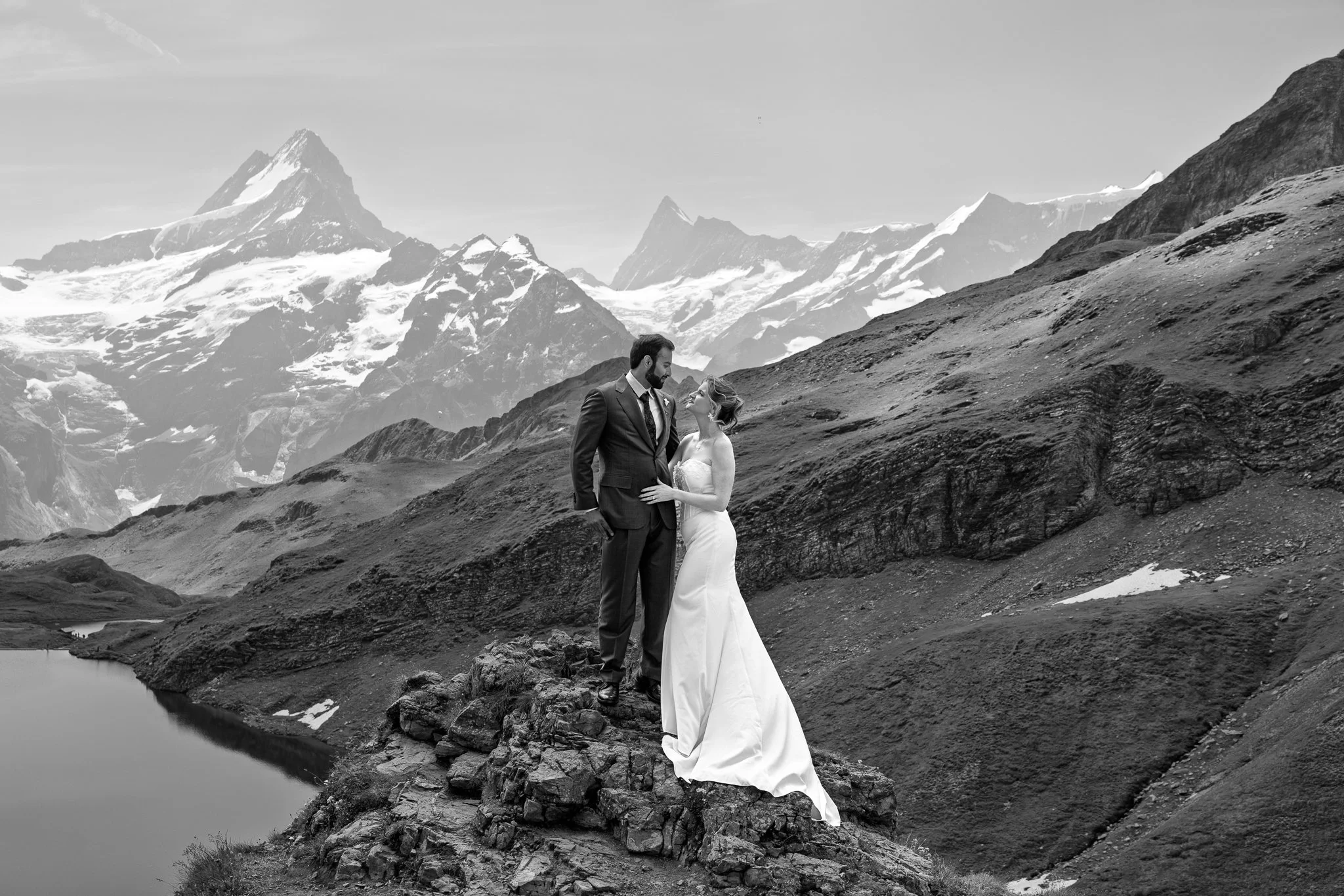 A black-and-white photograph of a bride and groom standing on a rocky ledge in a mountainous landscape. The bride is wearing a white wedding dress, and the groom is in a dark suit. Behind them are snow-capped mountains and a body of water.
