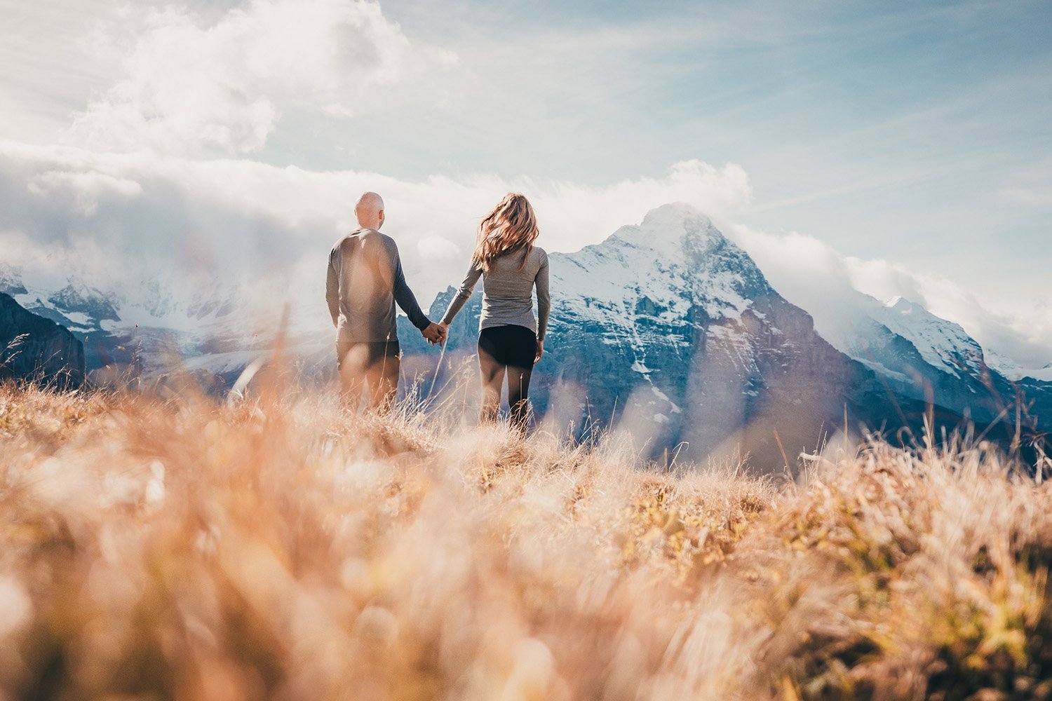 A couple holding hands and walking through a field of tall grass towards snow-capped mountains under a cloudy sky.