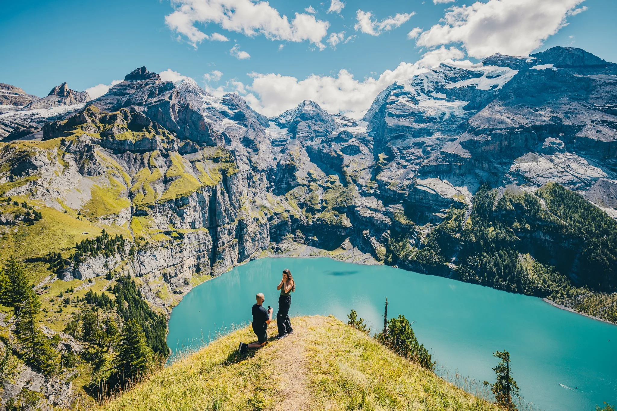 A man proposing marriage to a woman in front of a mountain lake surrounded by mountains and greenery. Lake Oeschinen