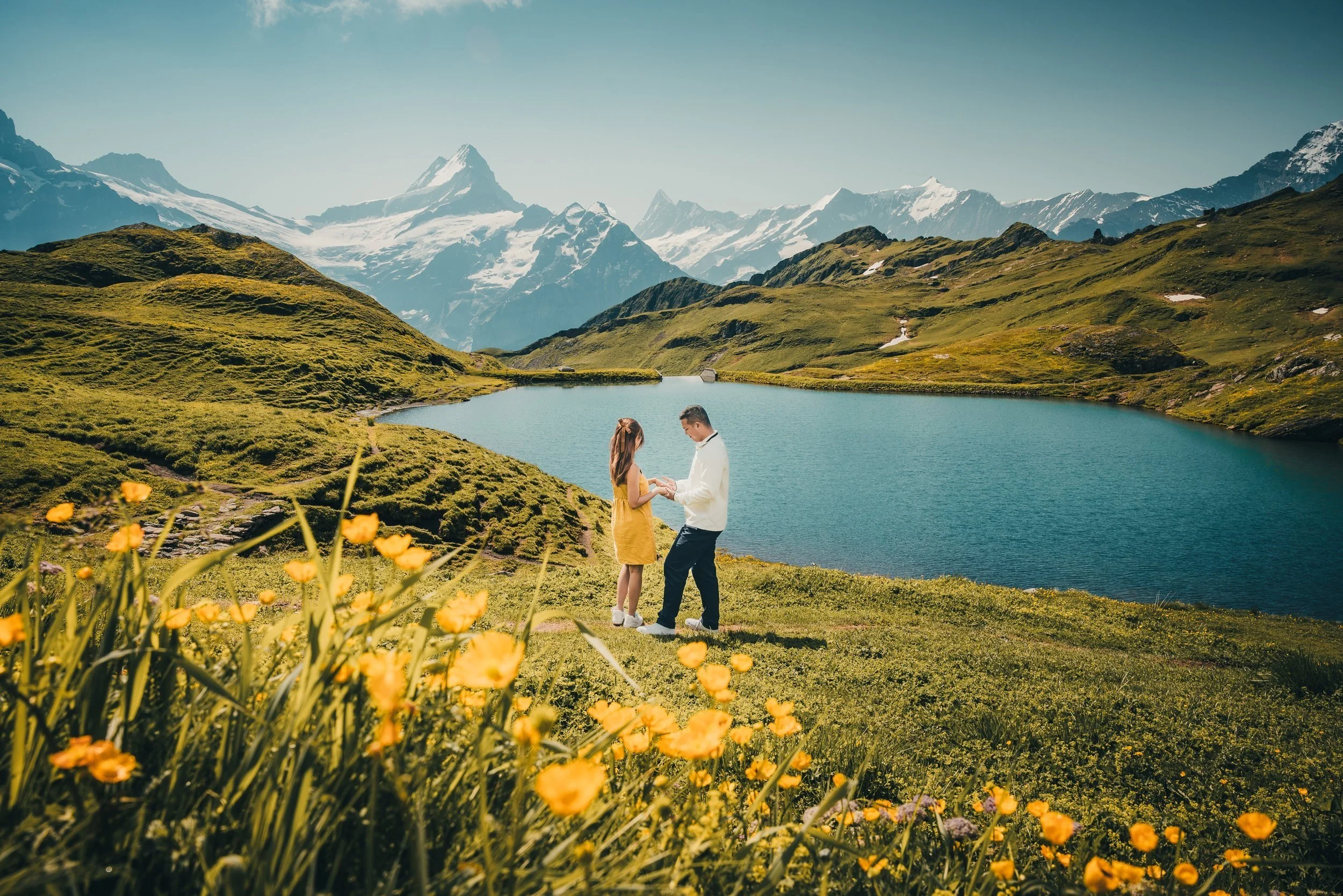 A couple standing by a lake in a mountainous landscape with snow-capped peaks in the background. The woman is wearing a yellow dress and the man is in a white shirt and dark pants. They are holding hands and looking at each other, surrounded by green