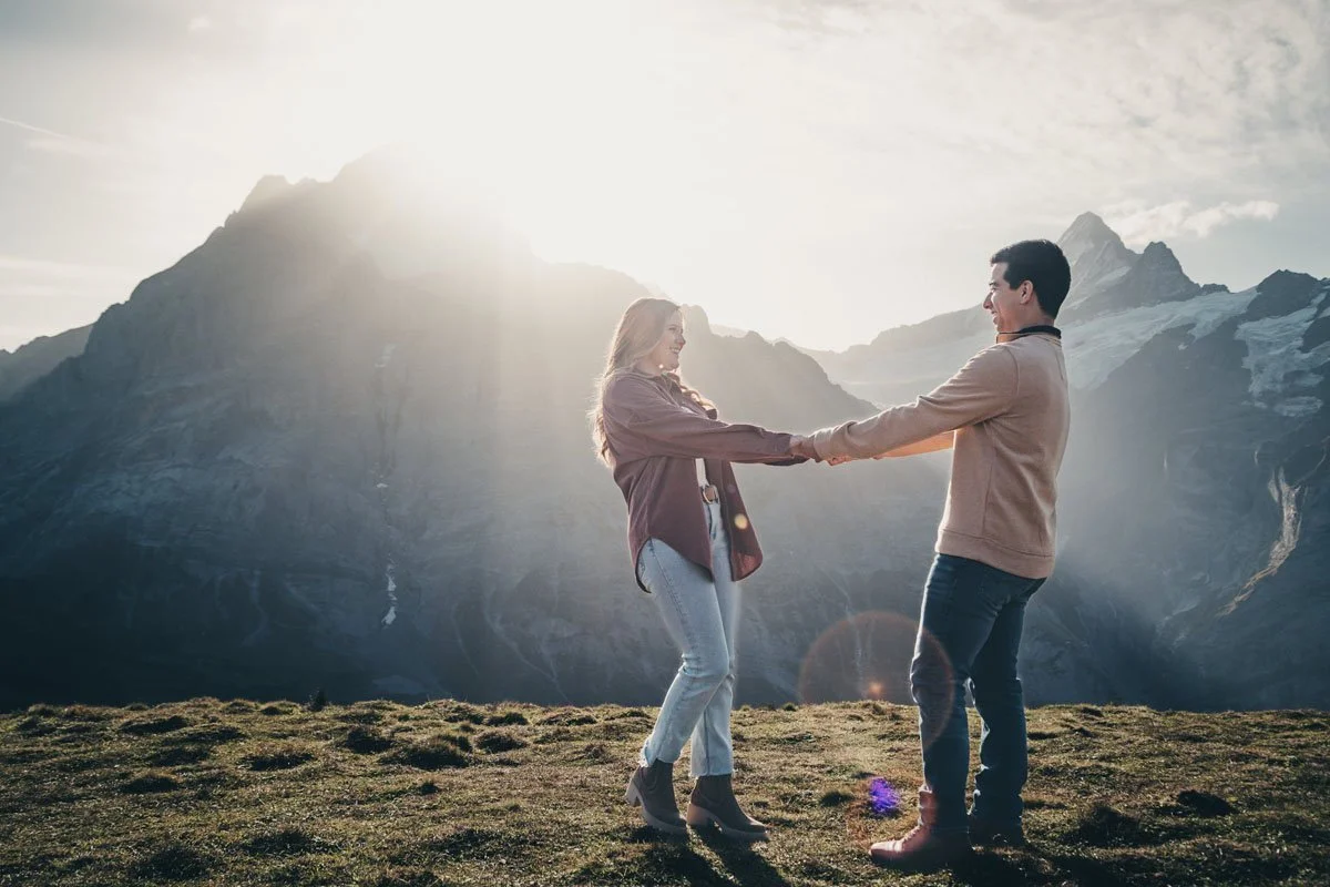 A man and woman holding hands and smiling at each other in a mountainous outdoor landscape at sunset.