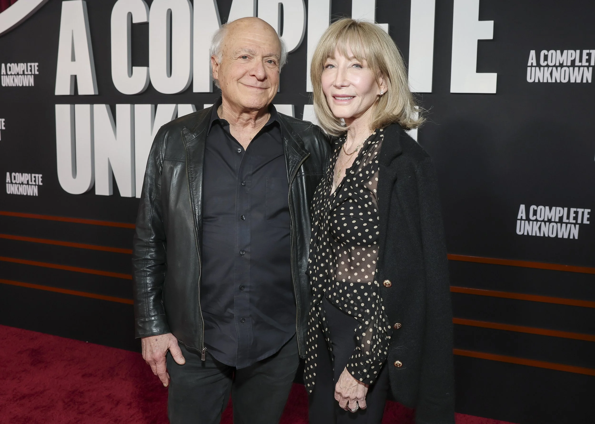 A man and woman standing on a red carpet in front of a black backdrop with white text. The man is wearing a black leather jacket over a black shirt and the woman is wearing a black polka dot blouse and a black cardigan. Bob Bookman.