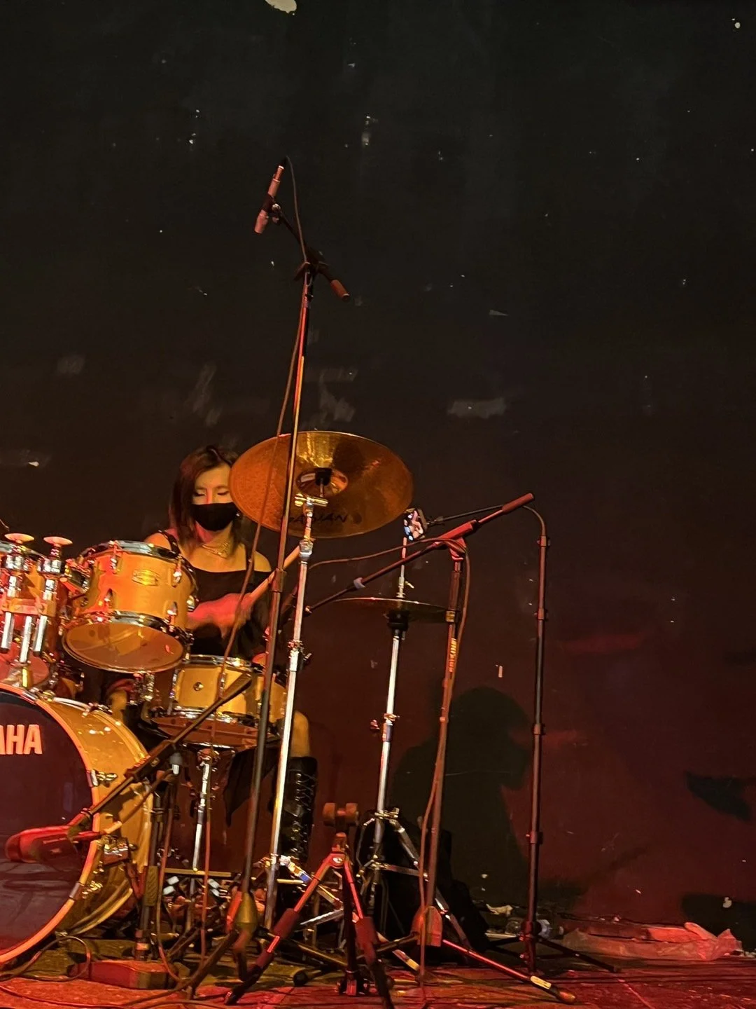 A Non binary drummer sitting behind a yellow drum set on a stage with microphones, wearing a black face mask, under stage lighting.