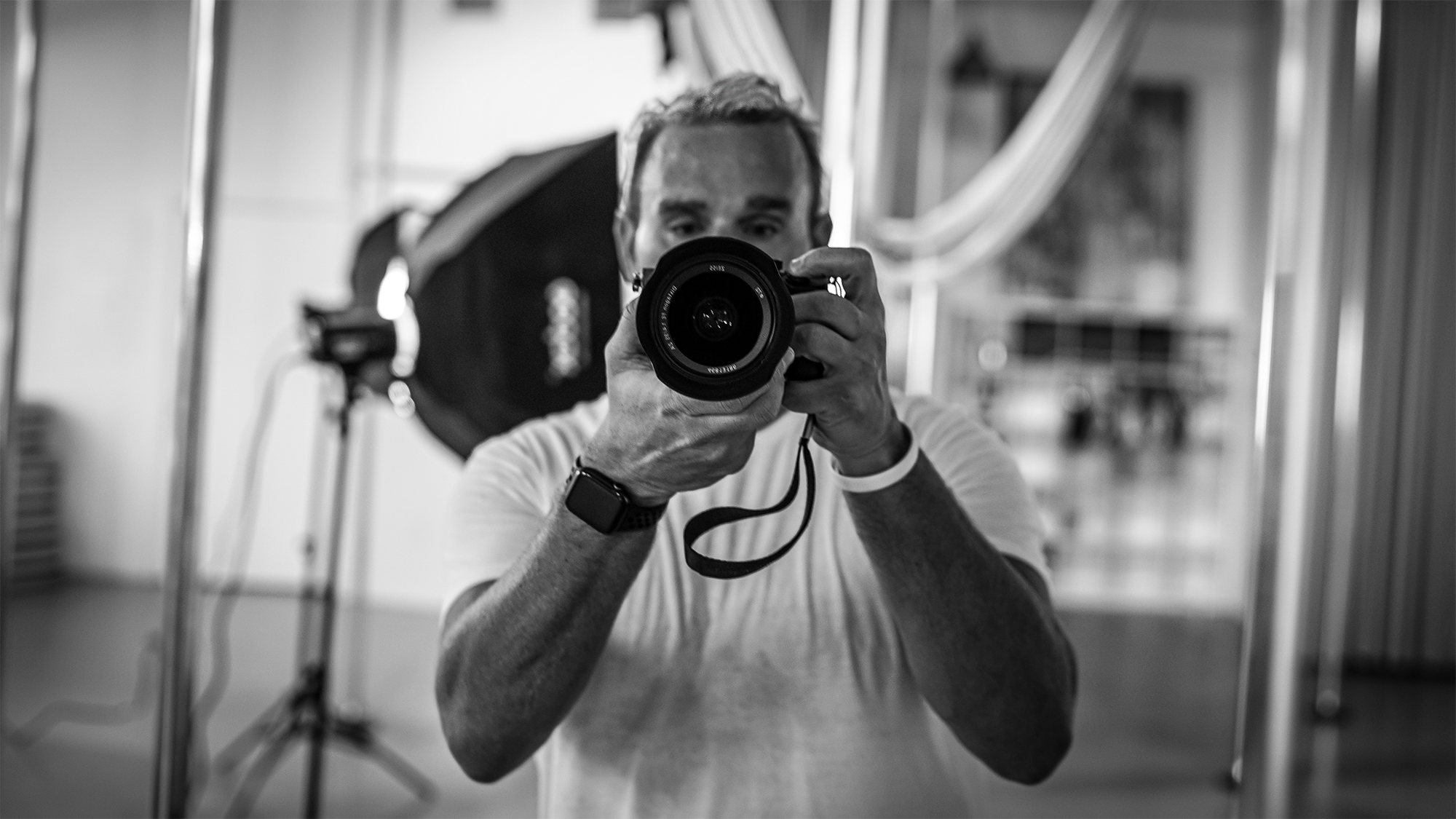 A man taking a photo with a camera, reflected in a mirror, in black and white. The man is holding a camera close to his face, with a background that appears to be a room with some furniture and a draped curtain.