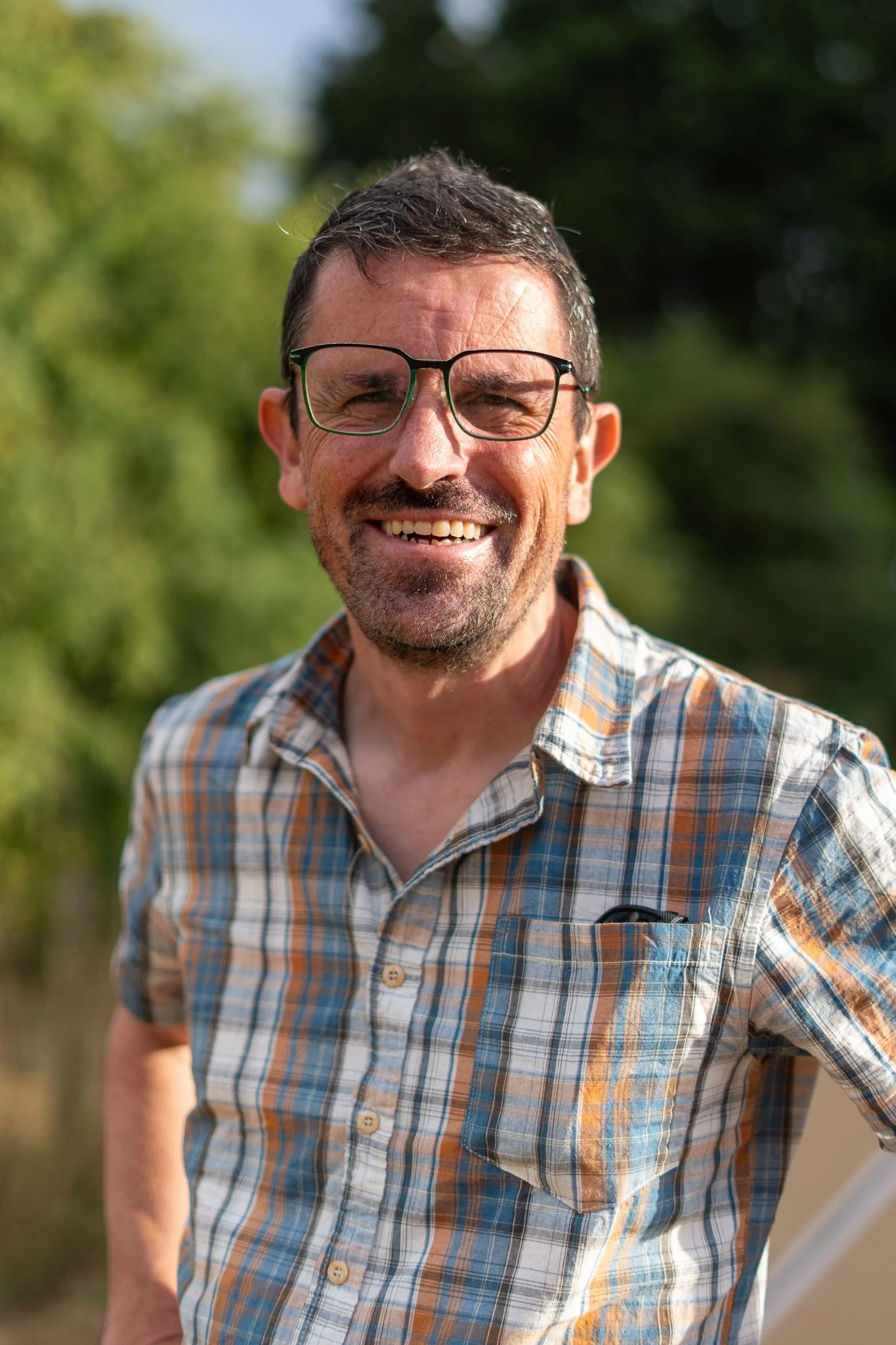 A smiling middle-aged man with glasses and a beard, wearing a plaid shirt, outdoors with blurred greenery in the background.