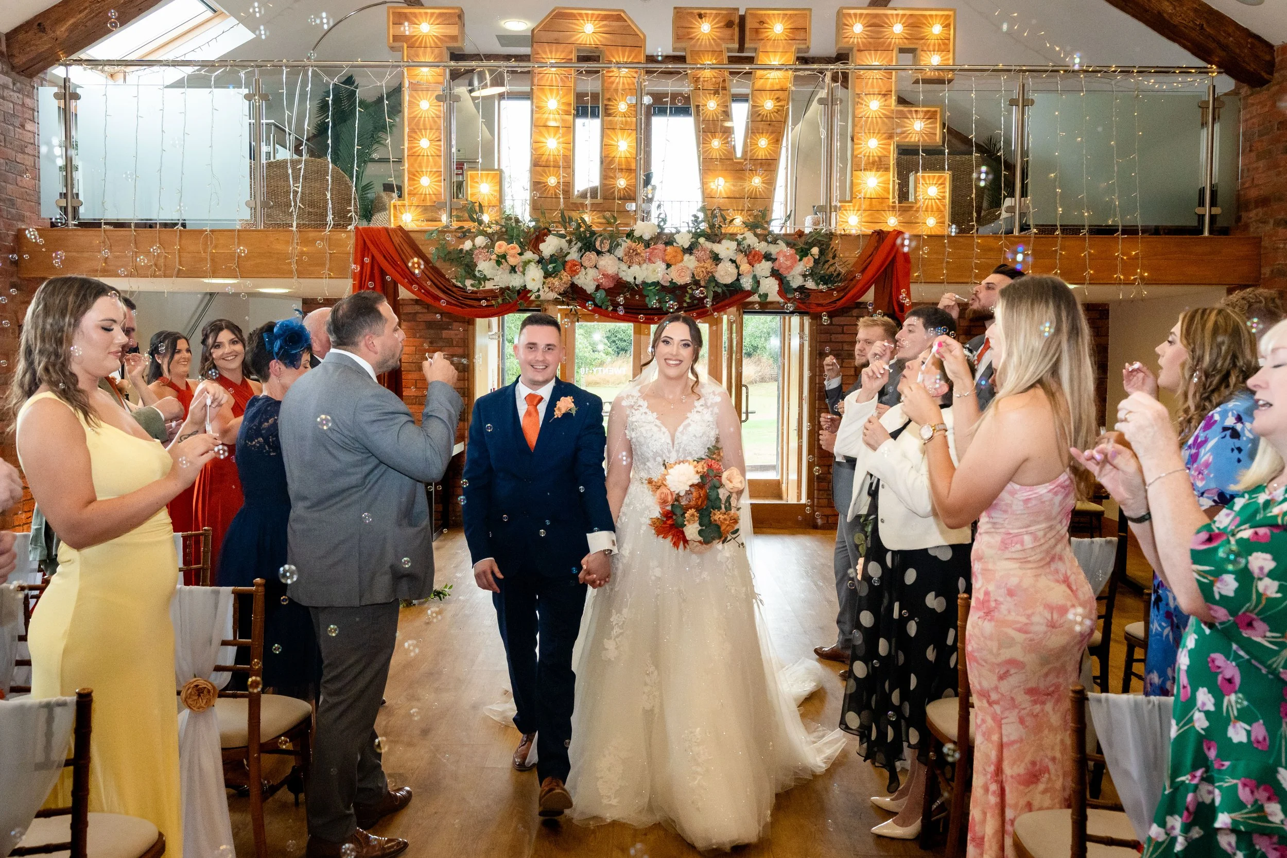 A bride and groom standing hand in hand in the center of a wedding reception, surrounded by wedding guests, with large illuminated letters spelling 'LOVE' and floral decorations overhead, in a brick-walled indoor venue.