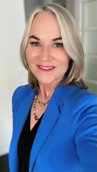 A woman with blonde hair smiling, wearing a blue blazer and a black top, standing indoors with a background of a door and white walls.