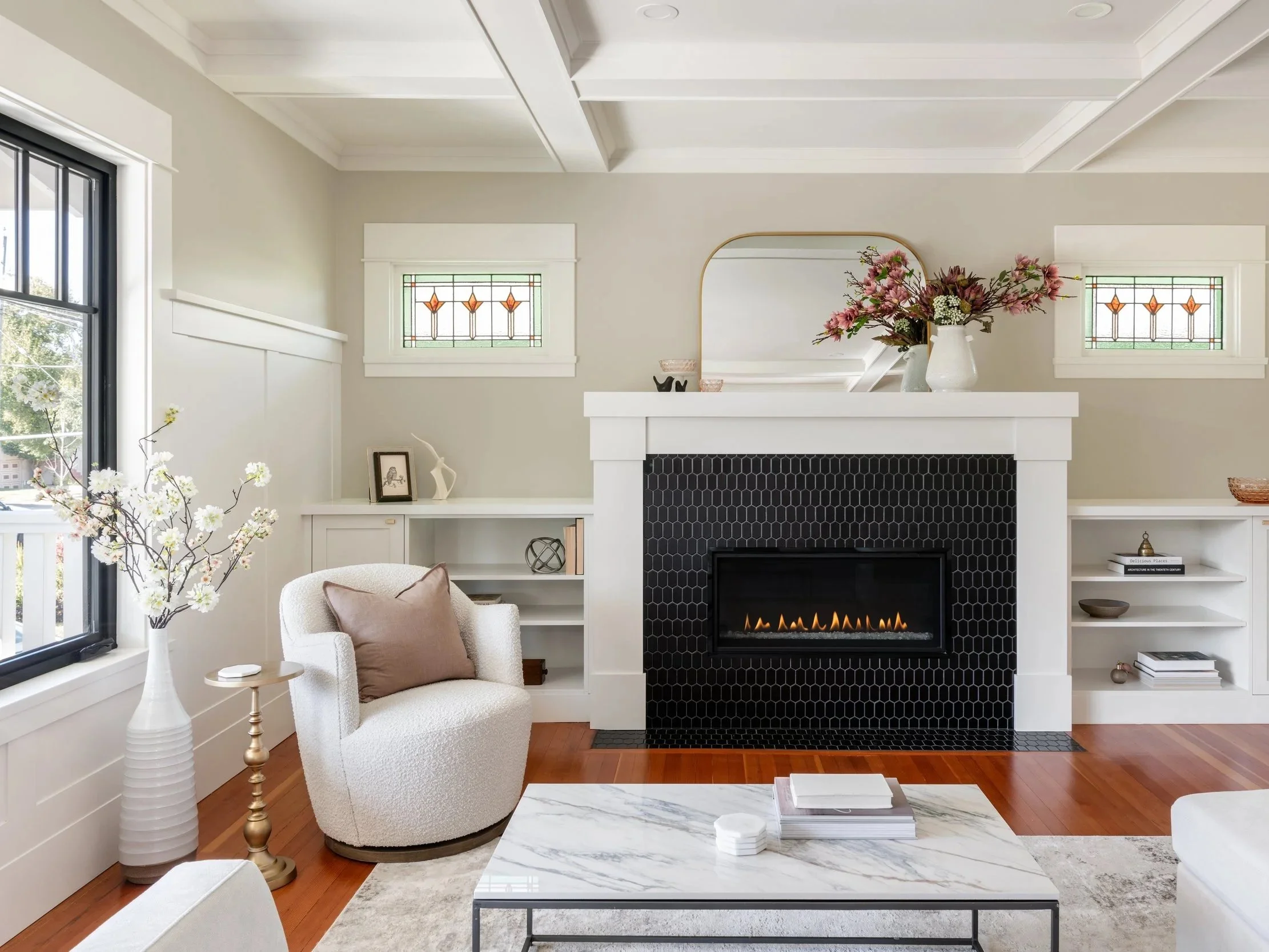 A cozy living room with a white armchair, a marble coffee table, a fireplace with black tiles, a large mirror, a vase with pink flowers, built-in shelves, and stained glass windows.