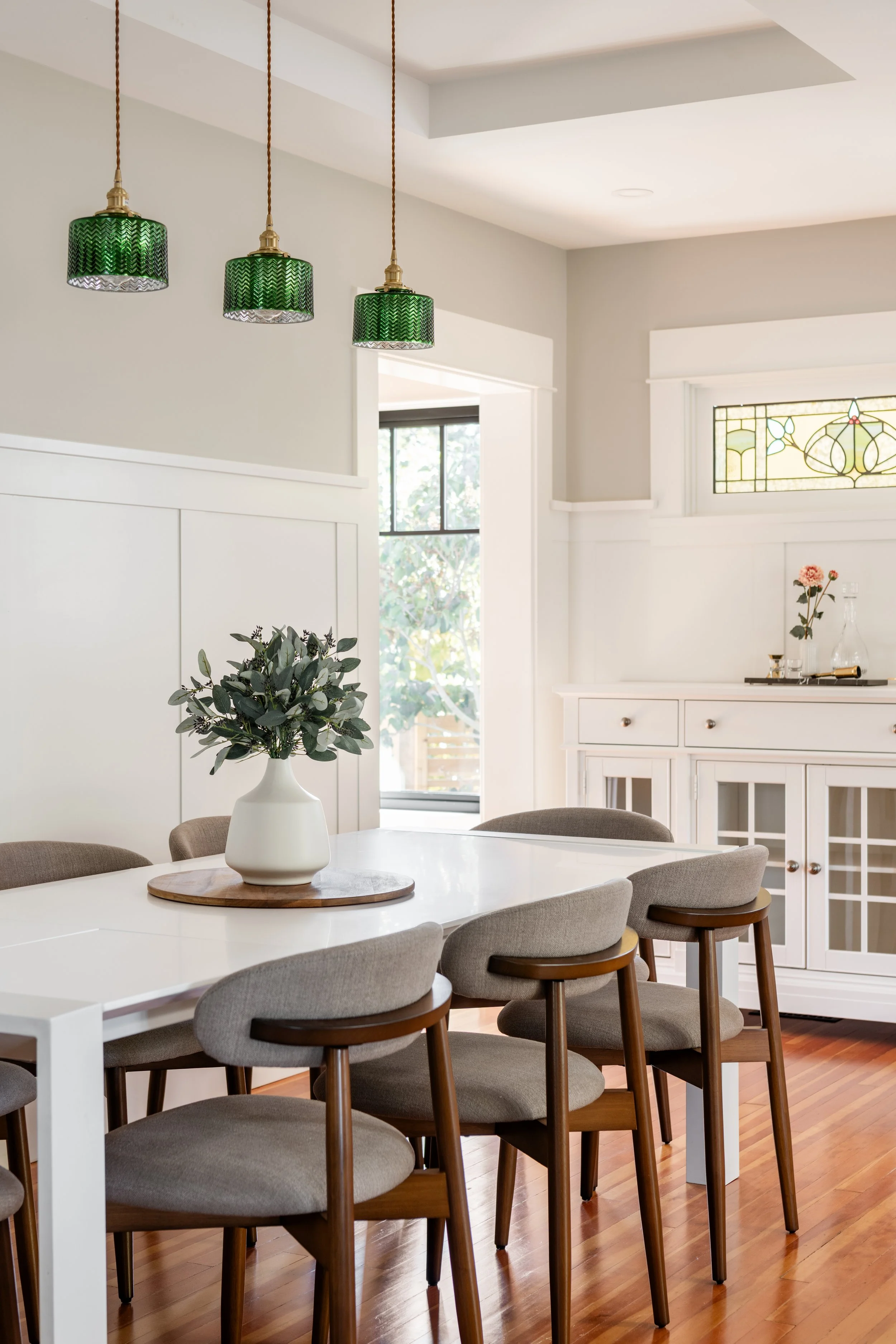 Dining room with white table, gray chairs, a flower vase, green pendant lights, and a white cabinet with glass doors.