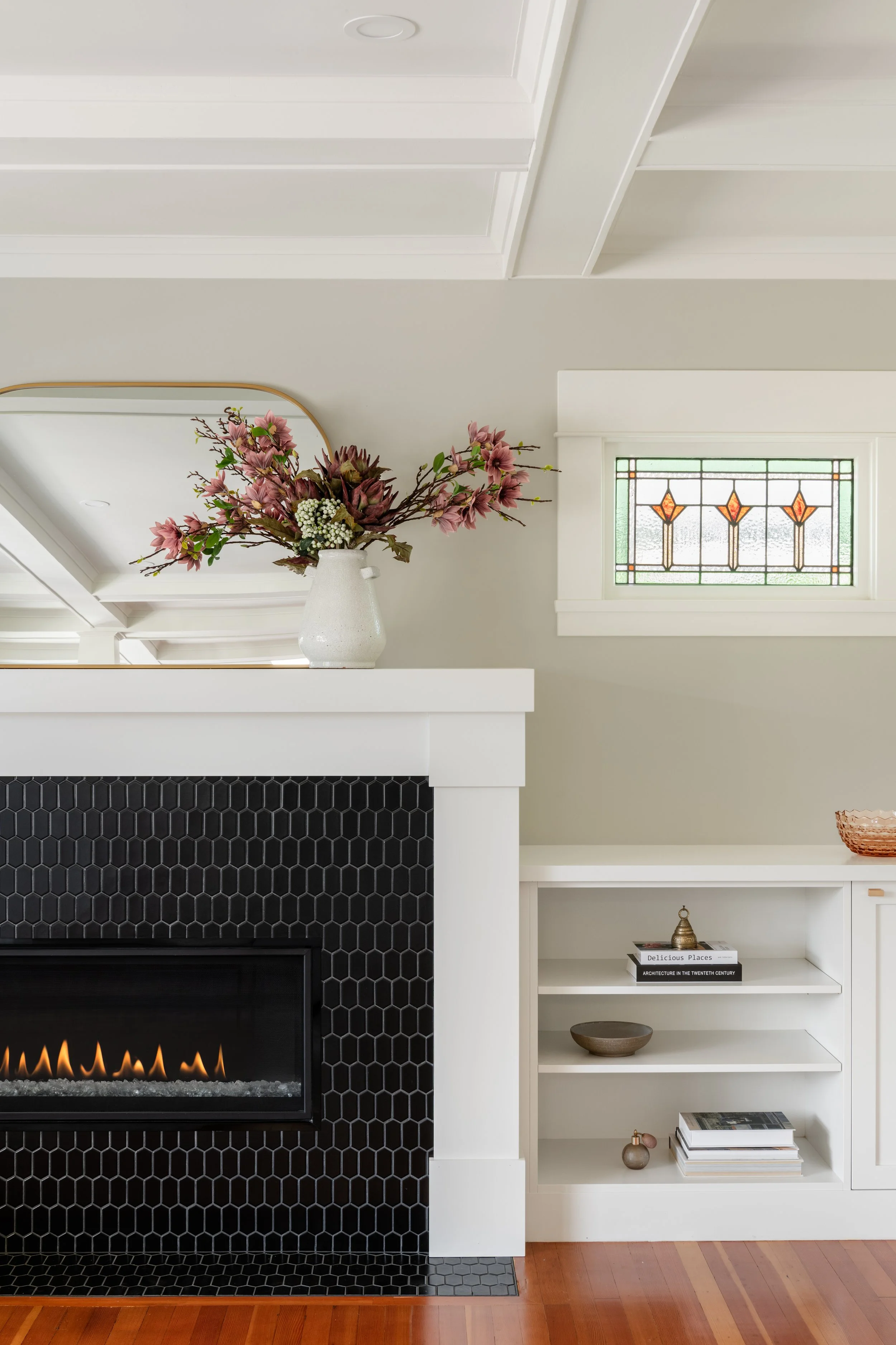 A cozy living room with a white fireplace surrounded by black hexagon tiles, a white built-in bookshelf with decorative items, a small stained glass window, and a large vase of pink flowers on top of the fireplace mantel.