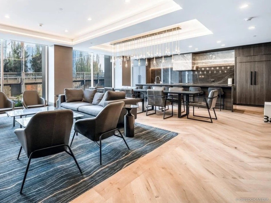 Modern lounge area with large windows, gray seating, a rug, a glass coffee table, and a kitchen island with bar stools in the background.