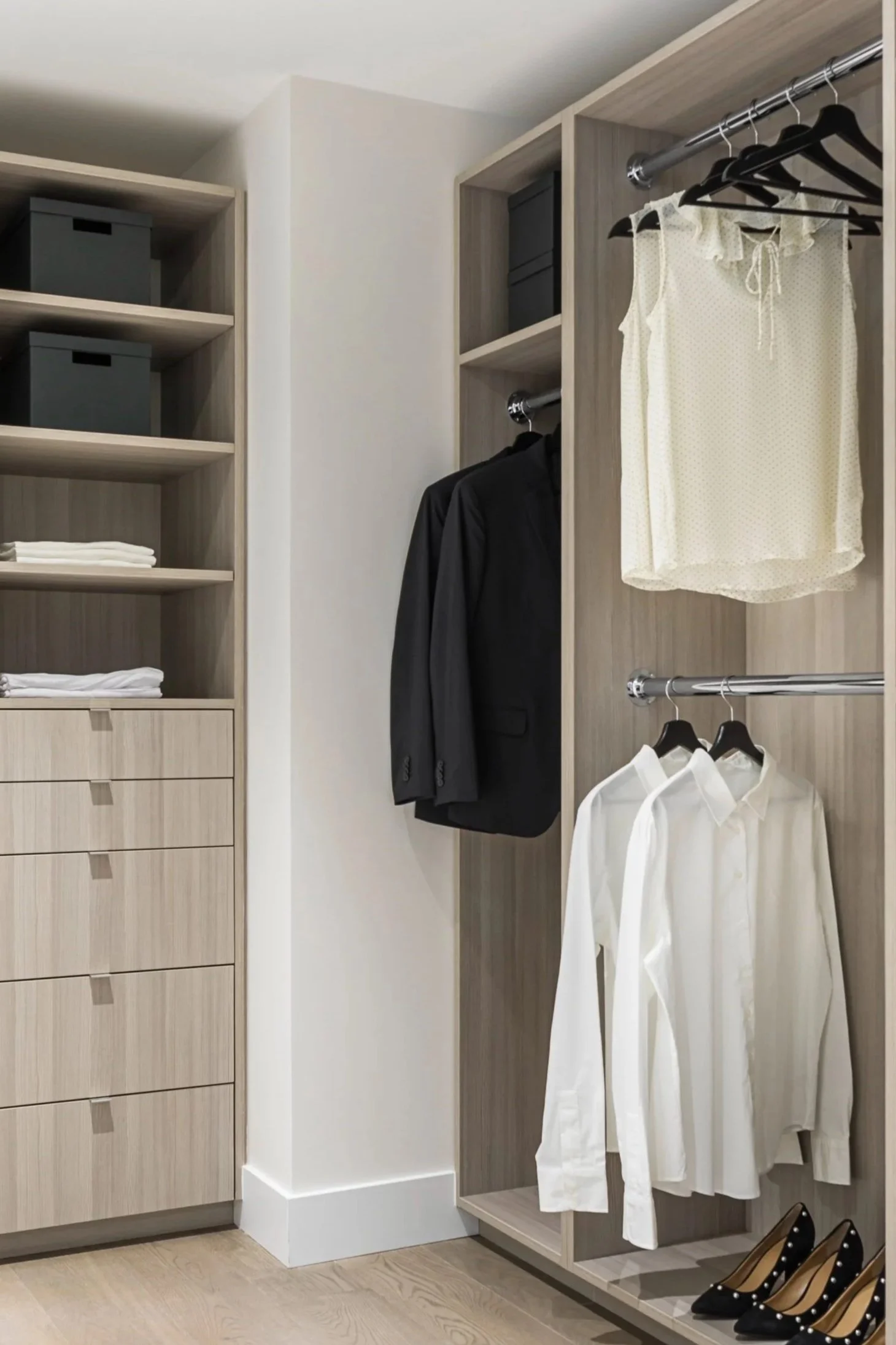 A walk-in closet with hanging clothing and storage compartments, including a black suit, white shirts, a cream-colored sleeveless top, black polka dot high heels, and organized shelves with fabric boxes and folded towels.
